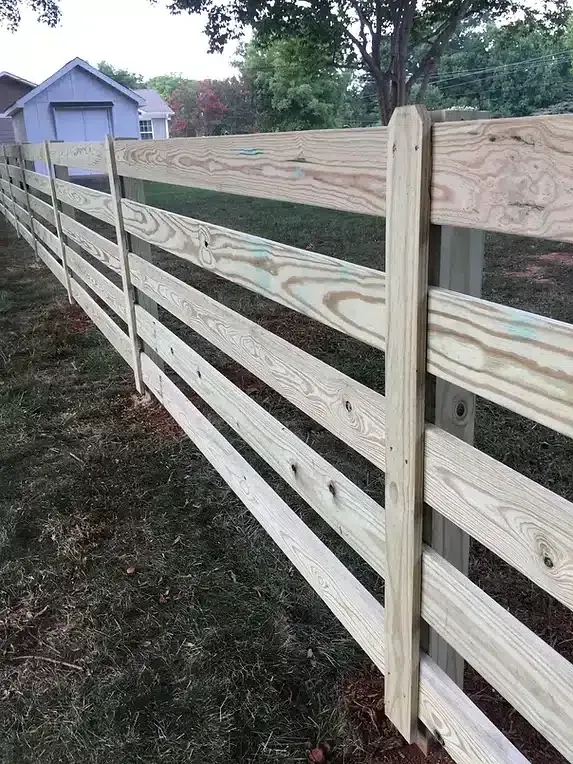 Wooden, four-rail fence in a grassy yard, with a small shed in the background.