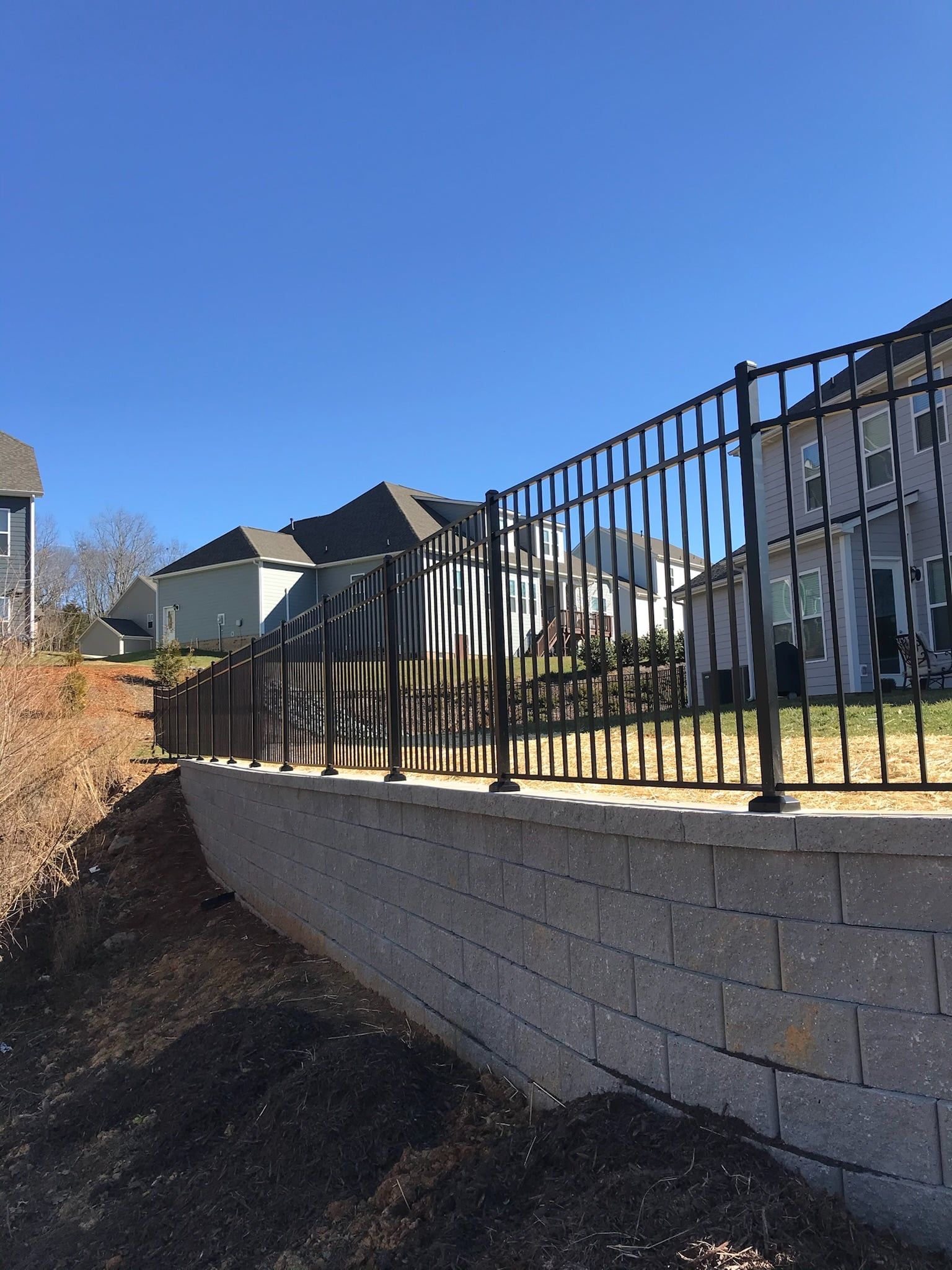 Black metal fence atop a gray retaining wall in front of houses on a sunny day.