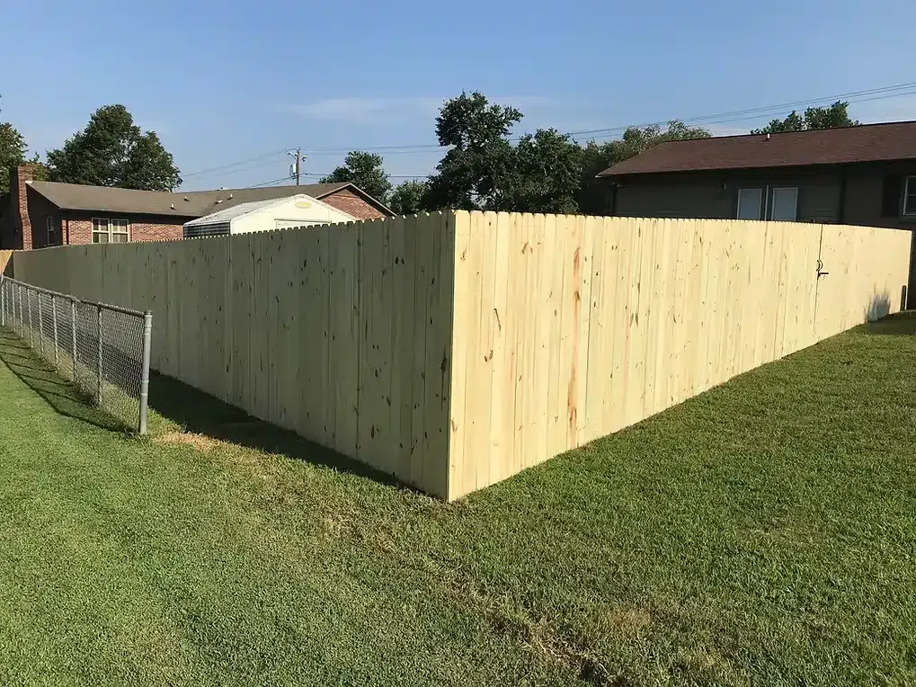 Wooden fence corner in a grassy yard, houses in the background on a sunny day.