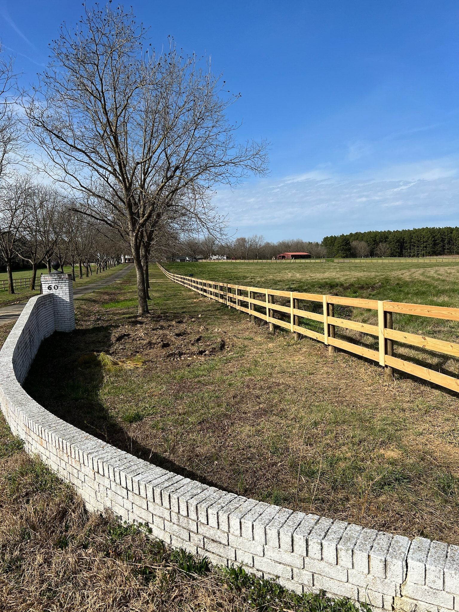 Curved white brick wall frames a tree, next to a wooden fence, with a field and forest in the background. Blue sky.