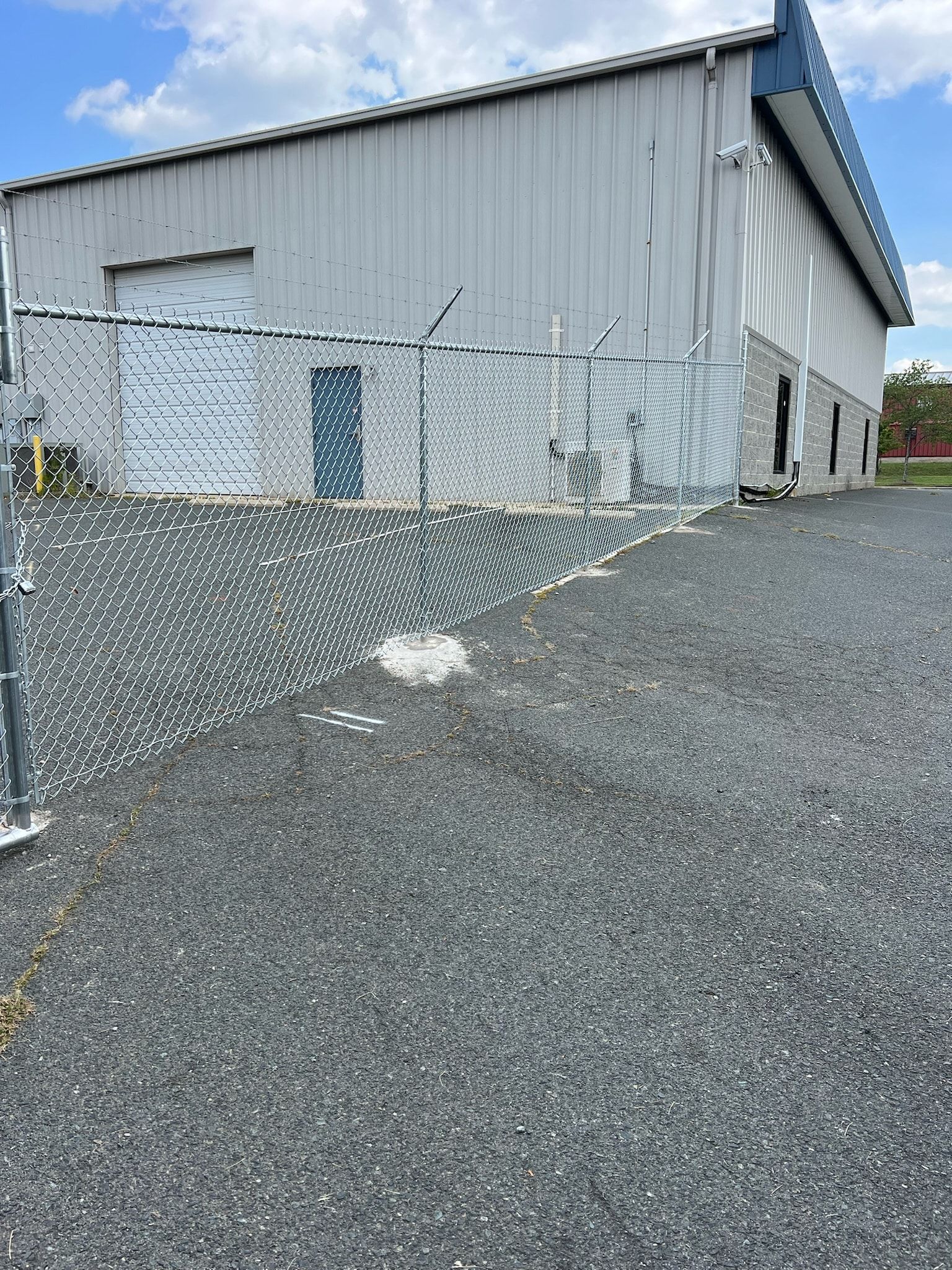 Chain-link fence in front of a gray industrial building on a gravel lot; sunny day.