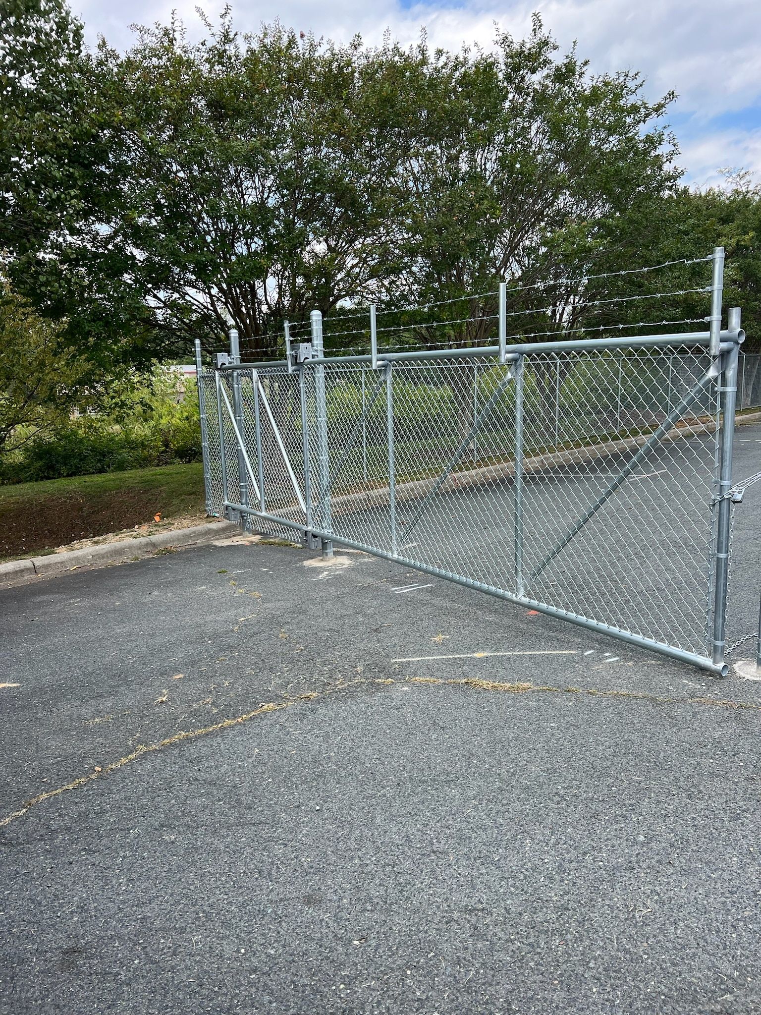 Metal chain link fence with an open gate on a gravel surface, trees in the background.