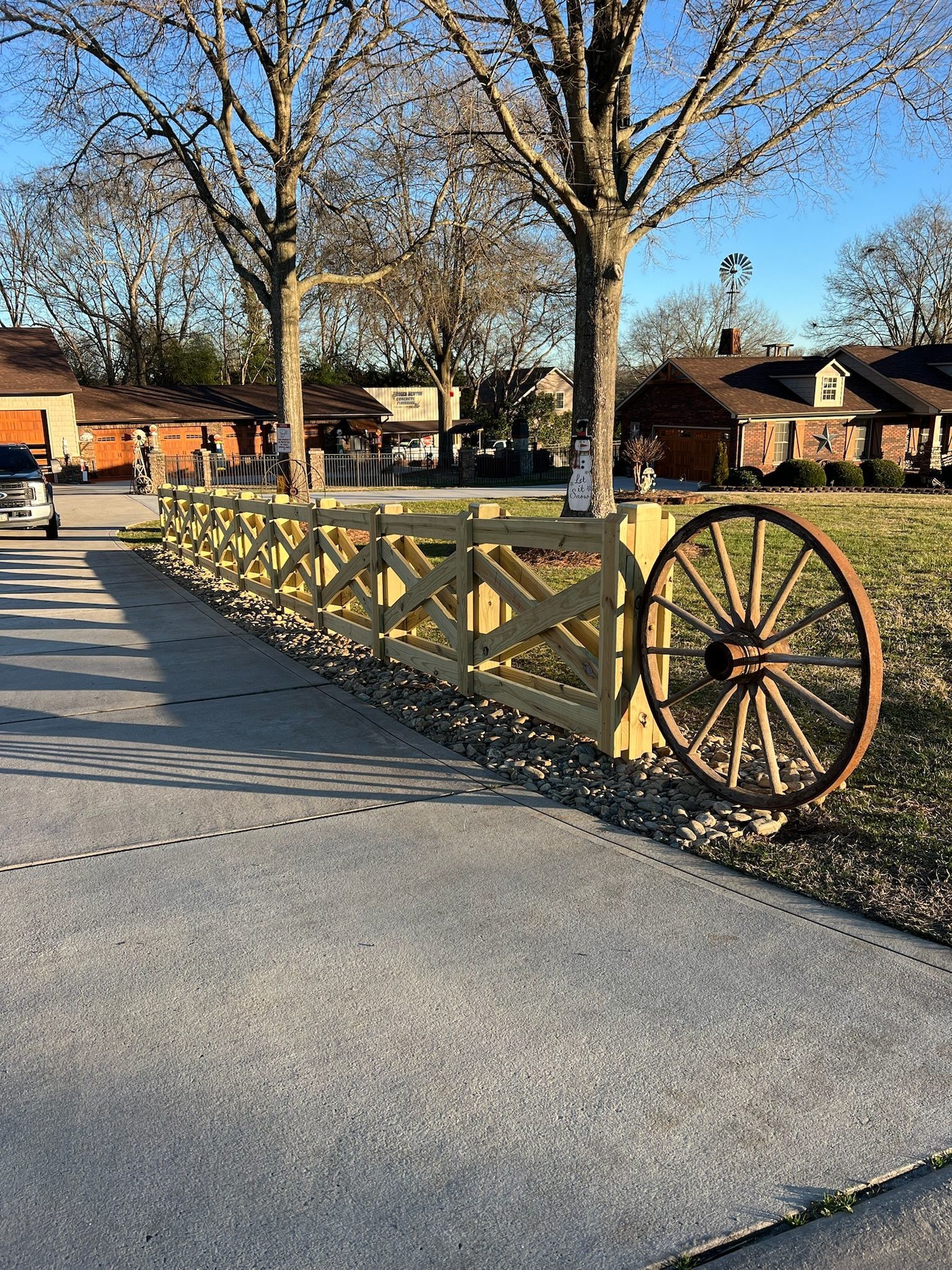 A wooden fence with a large wagon wheel decorates a yard in front of houses on a sunny day.