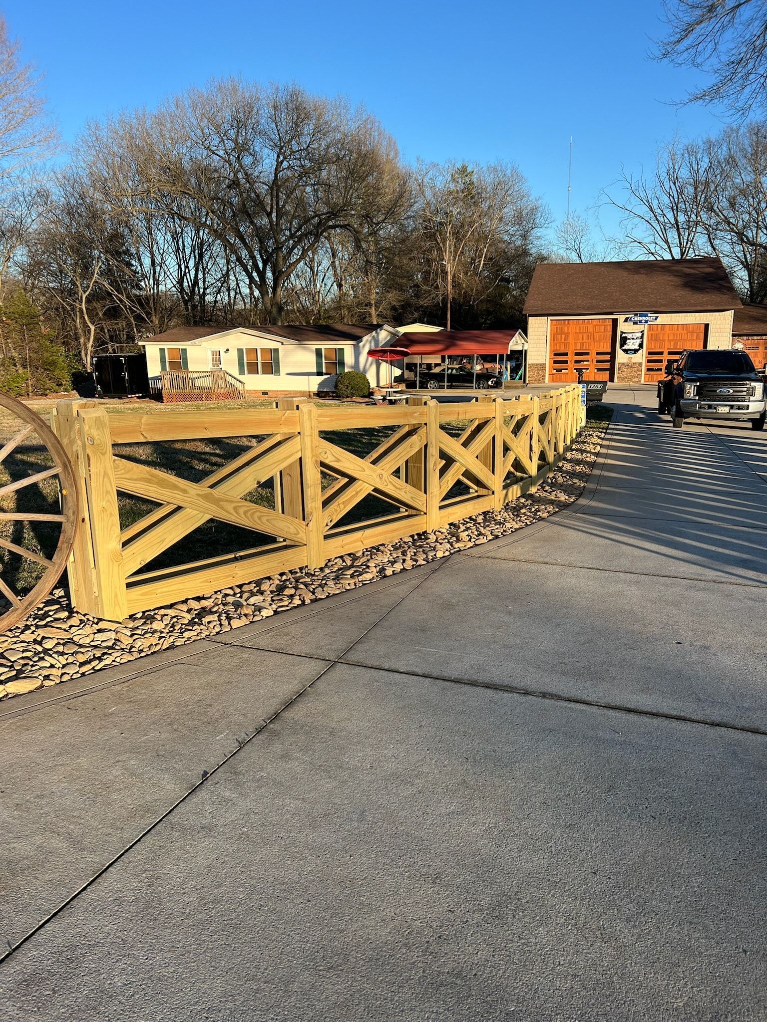 Wooden fence with crisscross design in front of a parking area, buildings and trees in the background.