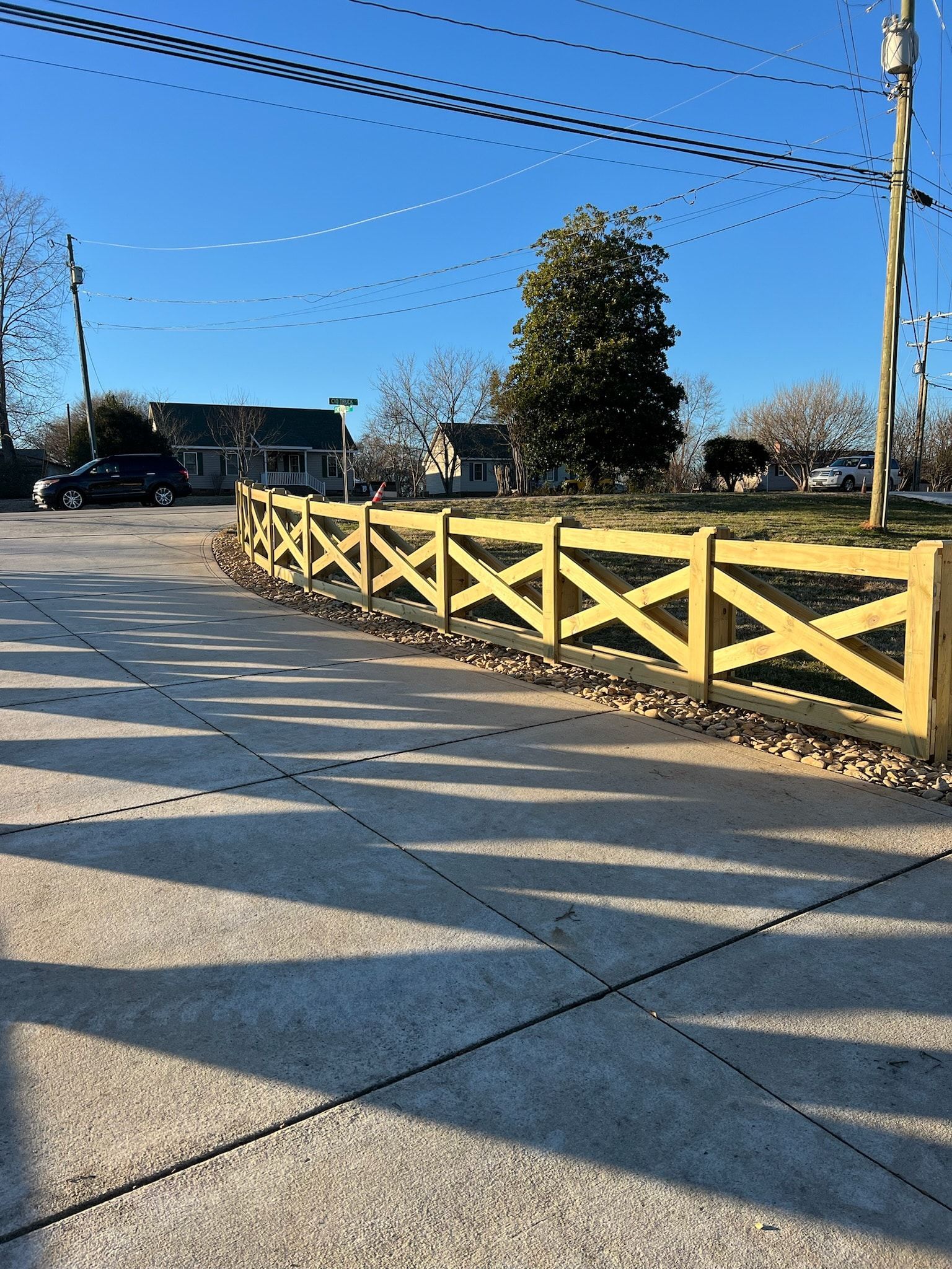 Yellow wooden fence with an X design bordering a concrete area and a residential street on a sunny day.