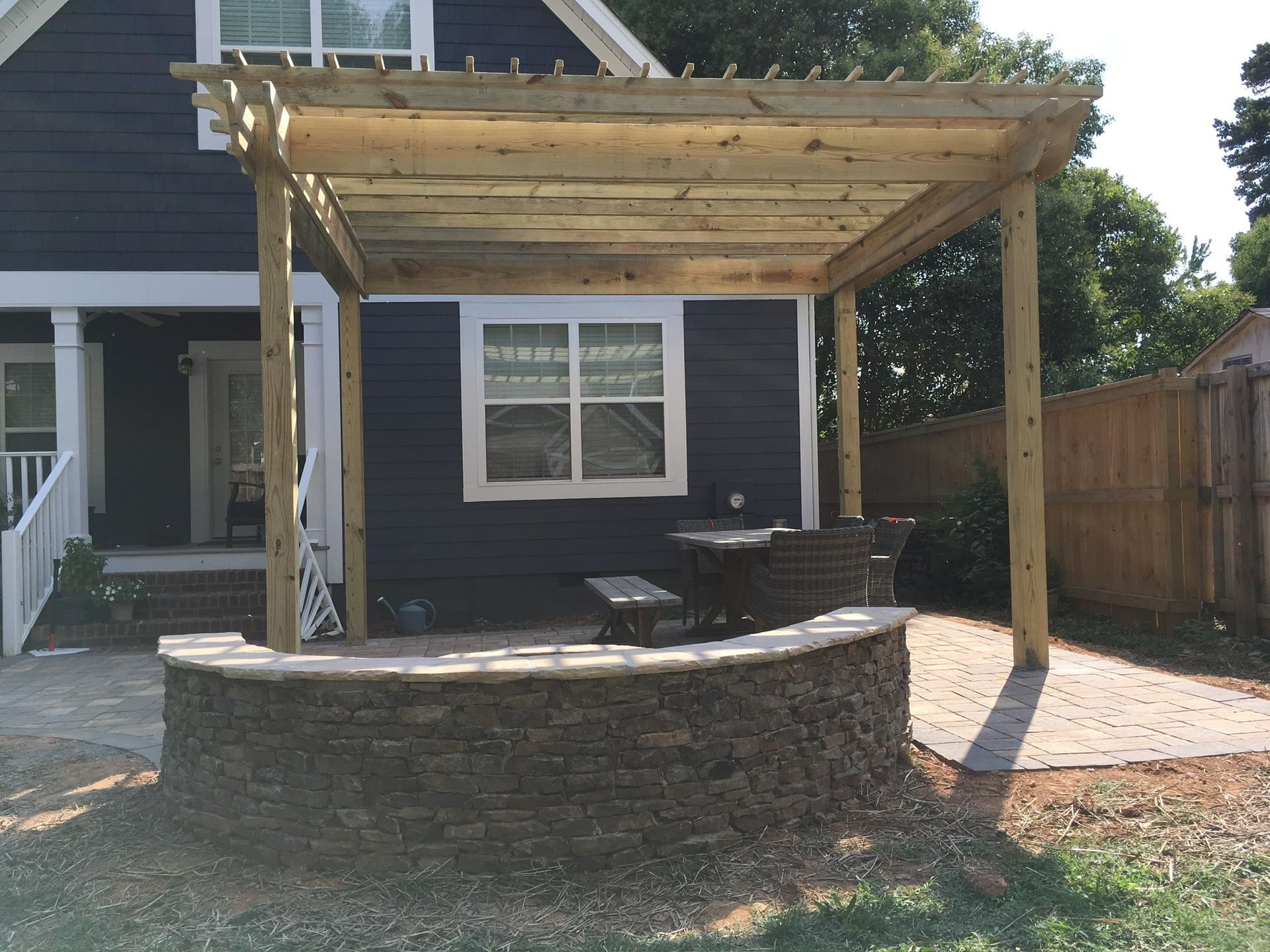 Wooden pergola over a stone-walled patio, beside a dark blue house.