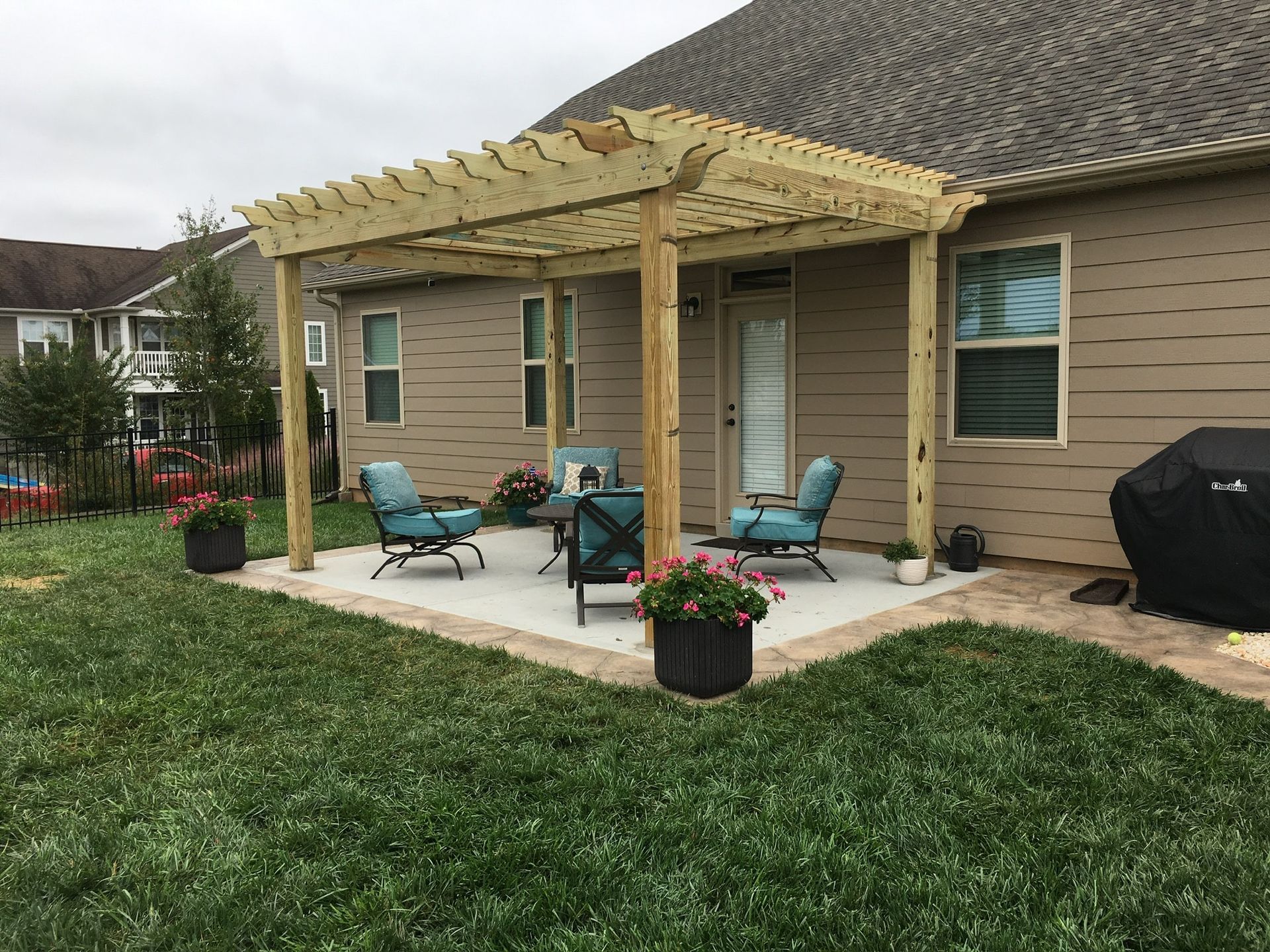 Backyard patio with pergola, seating, and flower pots, next to a house with tan siding and a grassy lawn.