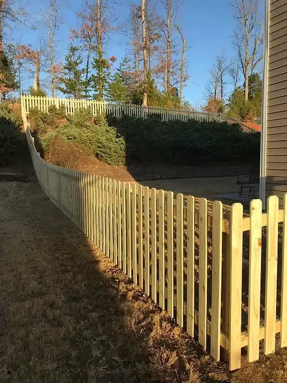 Wooden picket fence curving up a grassy hill, with trees and a blue sky in the background.