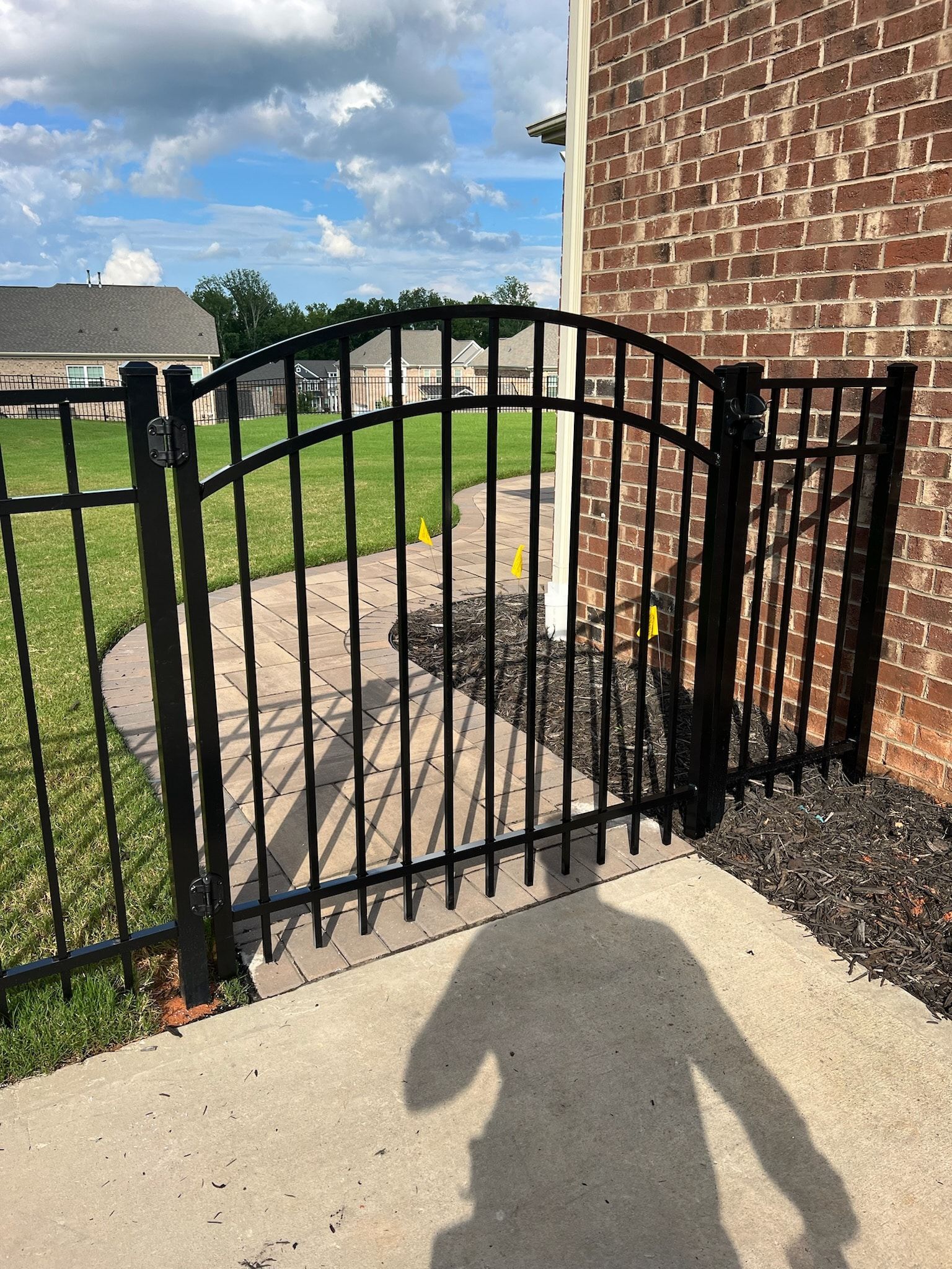 Black metal gate in a yard, leading to a stone path, beside a brick building. Sunny day.