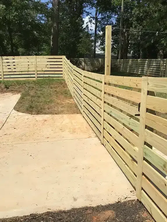 Wooden horizontal slat fence on a concrete surface, with grass and trees in the background.