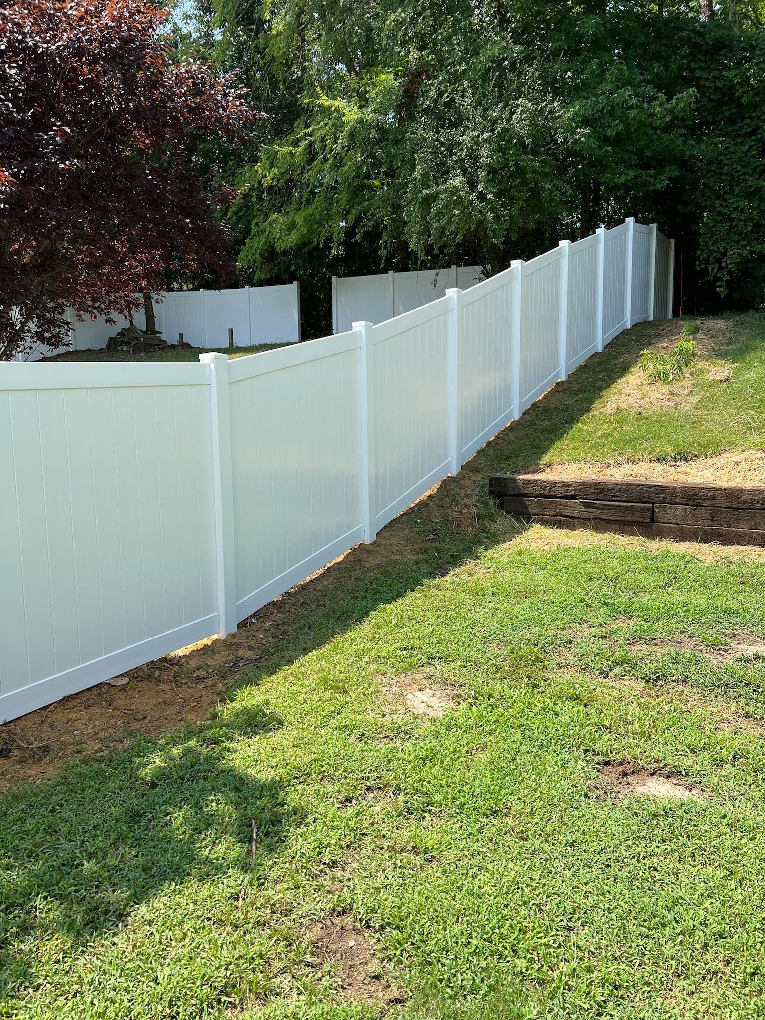 White vinyl fence on a grassy hill, with trees and a partial view of another fence in the background.