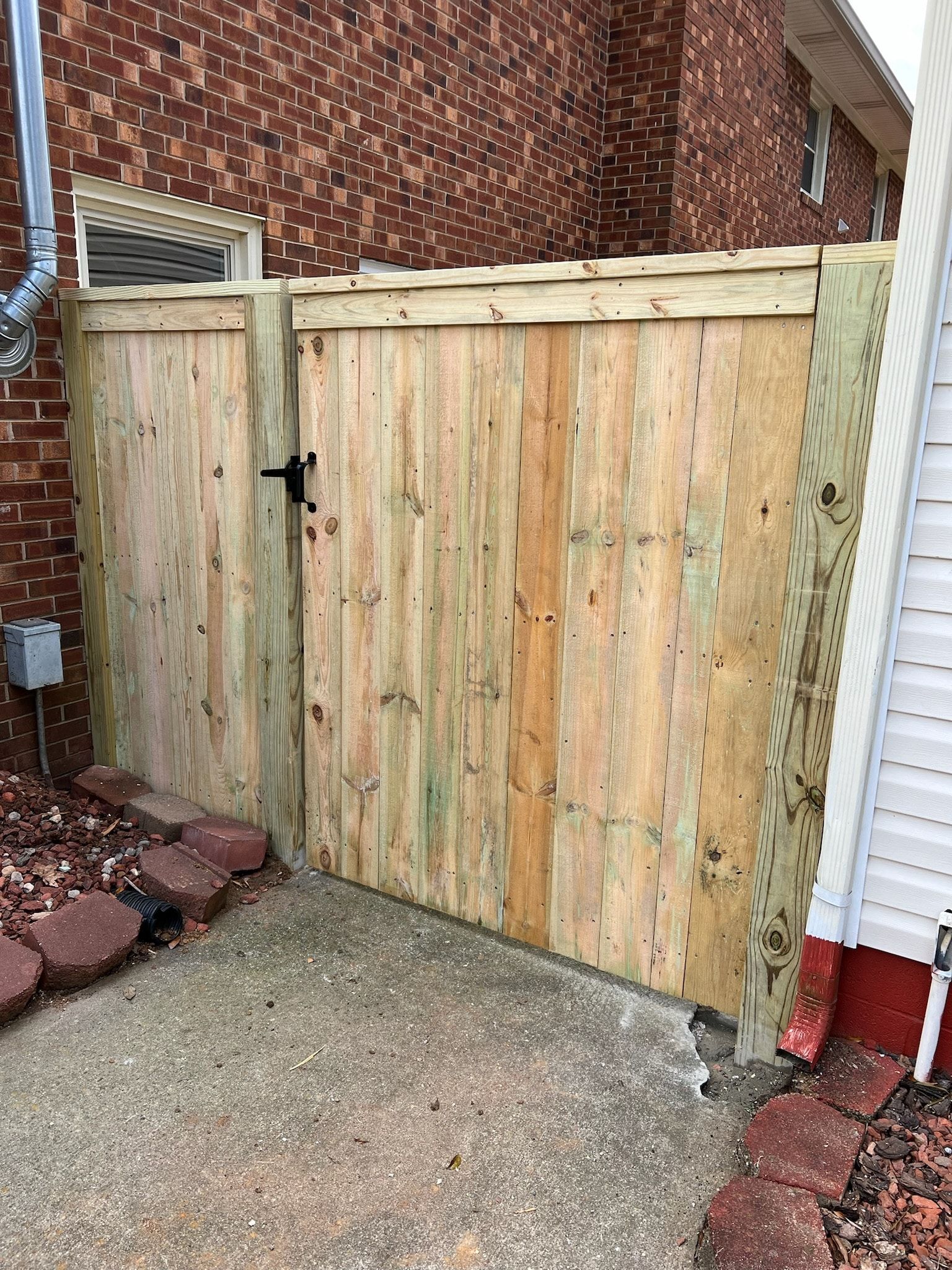 Wooden gate in a narrow concrete walkway, against a brick building and a white-sided house.