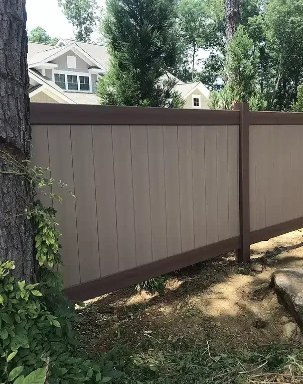 Tan and brown privacy fence in front of houses, with a tree on the left and foliage in the foreground.