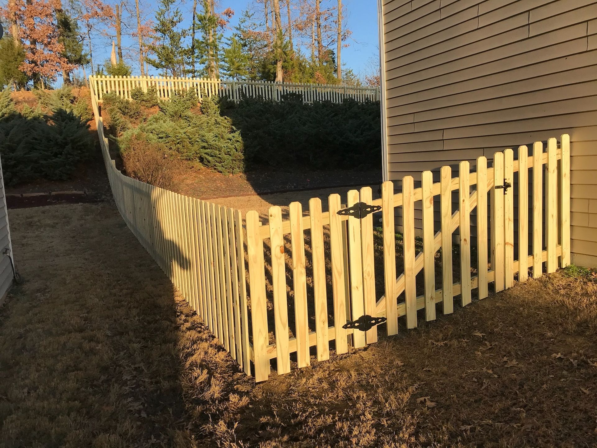 Wooden picket fence with gate bordering a yard with landscaping against a siding wall.