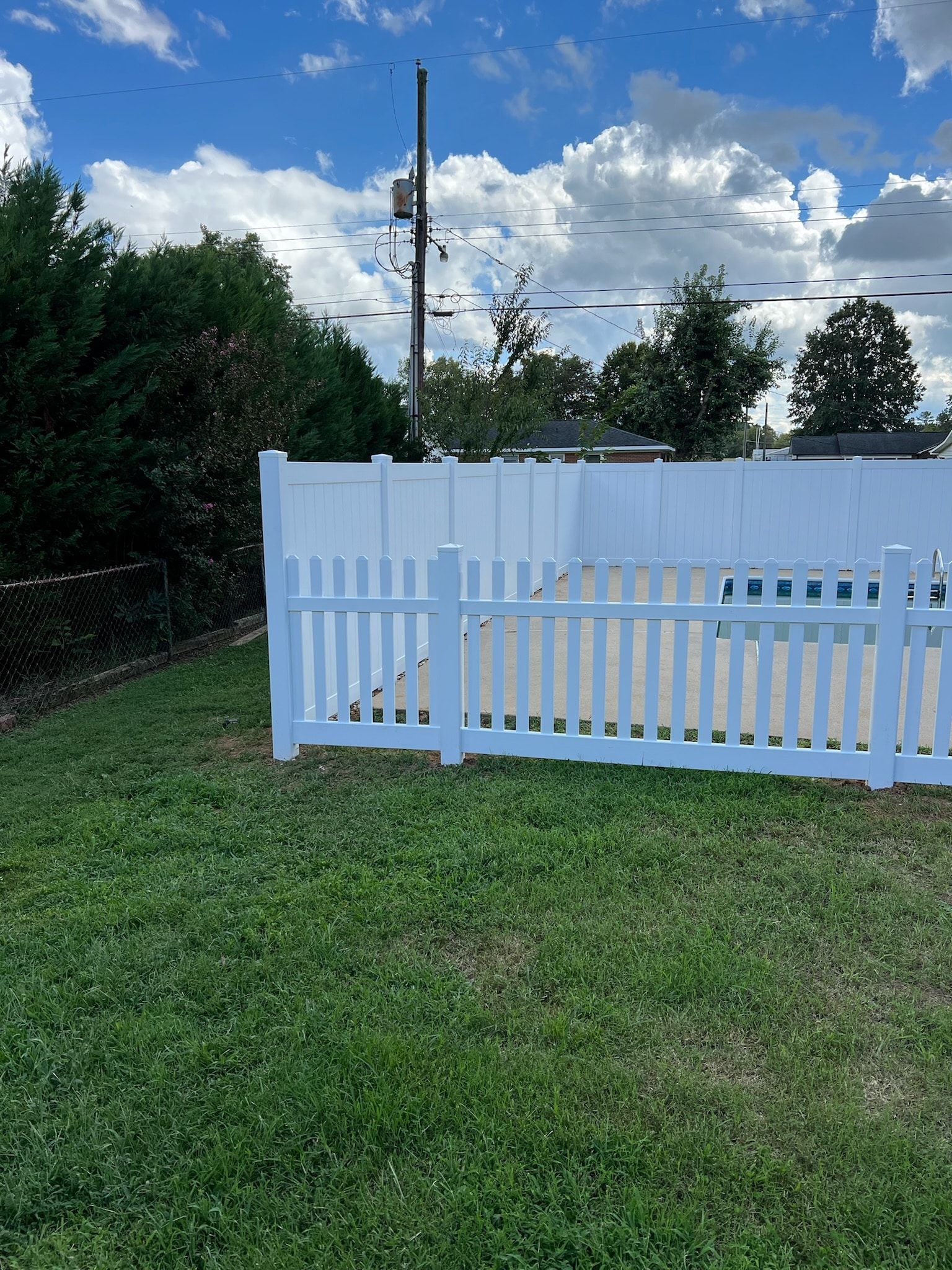 White picket fence in a grassy yard, trees in background, a utility pole, and a partly cloudy sky.