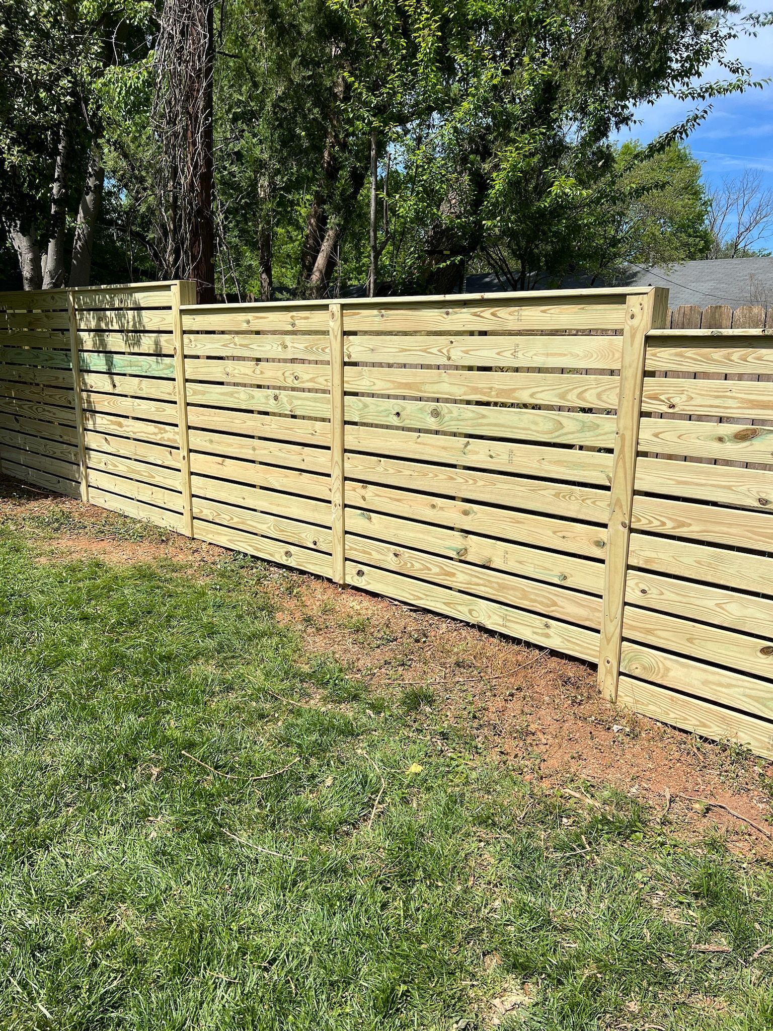 A horizontal wooden fence in a yard with green grass and trees in the background.