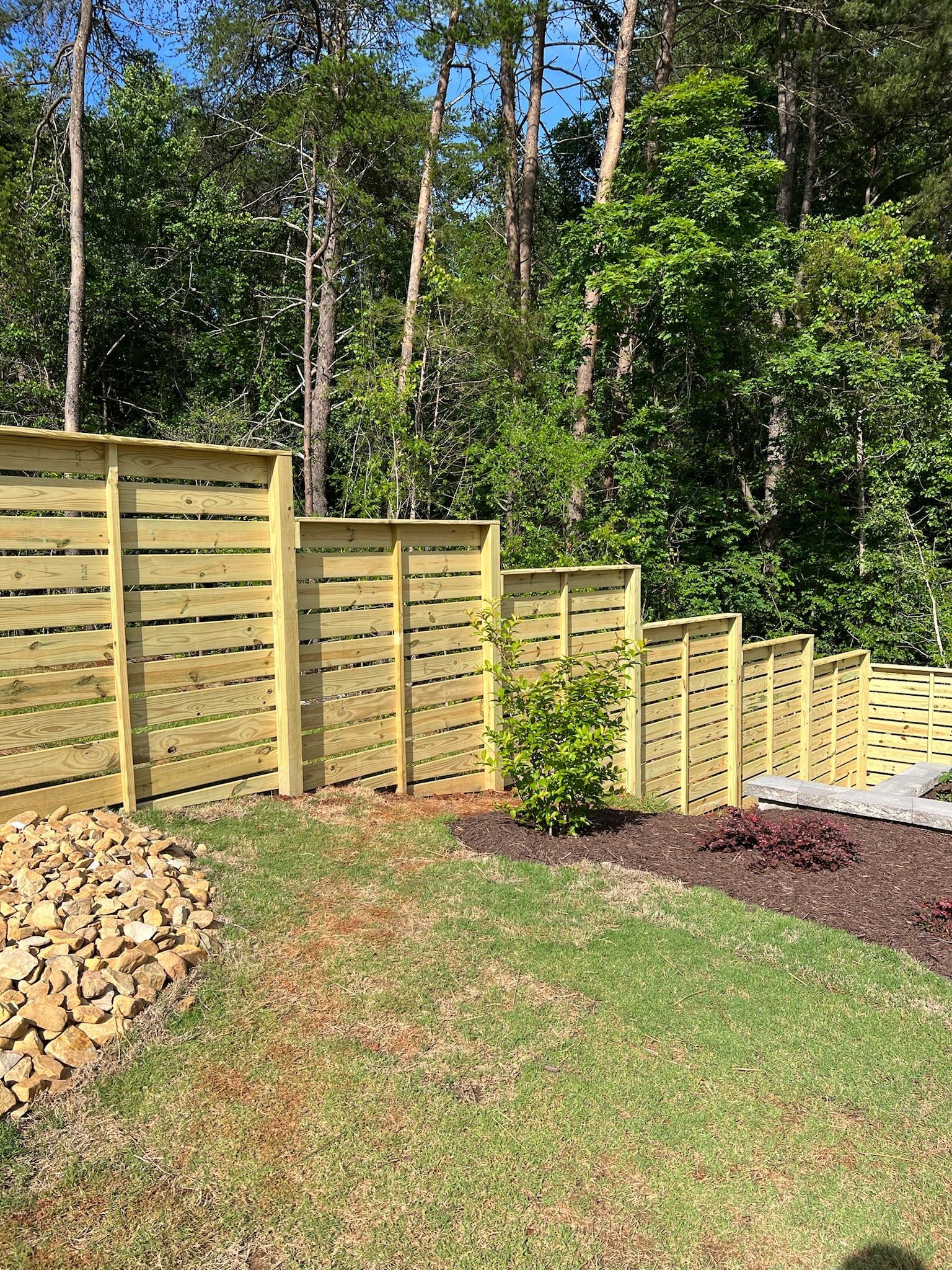 Wooden horizontal slat fence on a grassy hill, separating a yard from a wooded area.