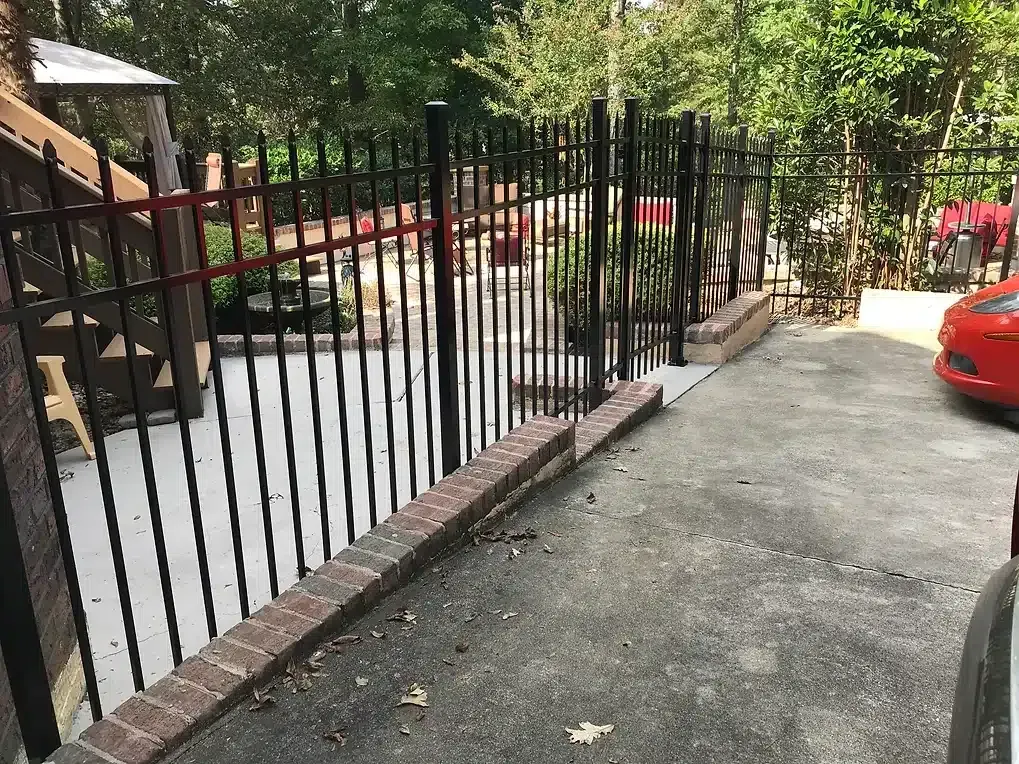 Black metal fence atop a brick wall bordering a driveway, with a red car parked at the right.