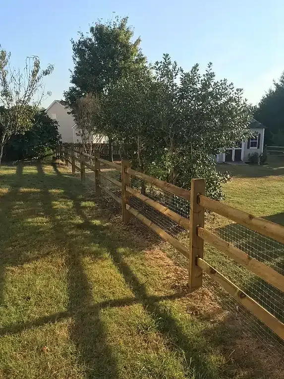 Wooden split-rail fence with wire mesh, running through grassy yard, trees and house in the background.