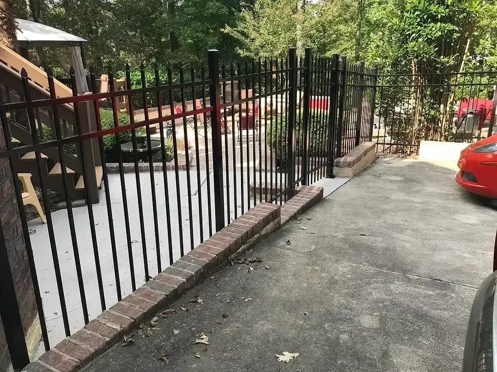 Black metal fence with brick base bordering a concrete driveway, red car parked to the right.