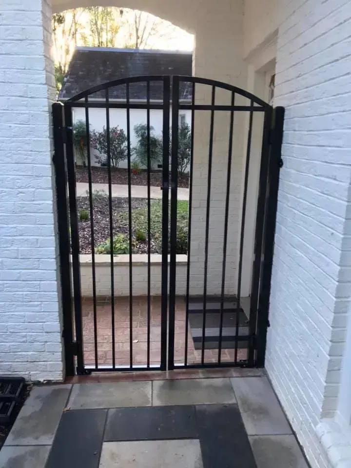 Black metal gate with vertical bars, set within a white brick entryway leading to a garden.