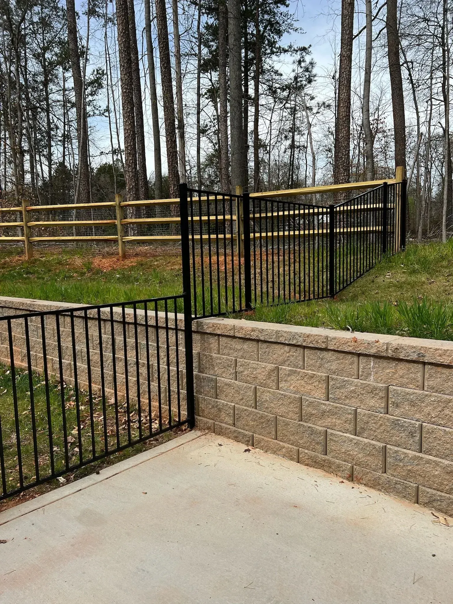 Black iron fence atop a retaining wall, with wooden fence behind, set in a grassy area with trees.
