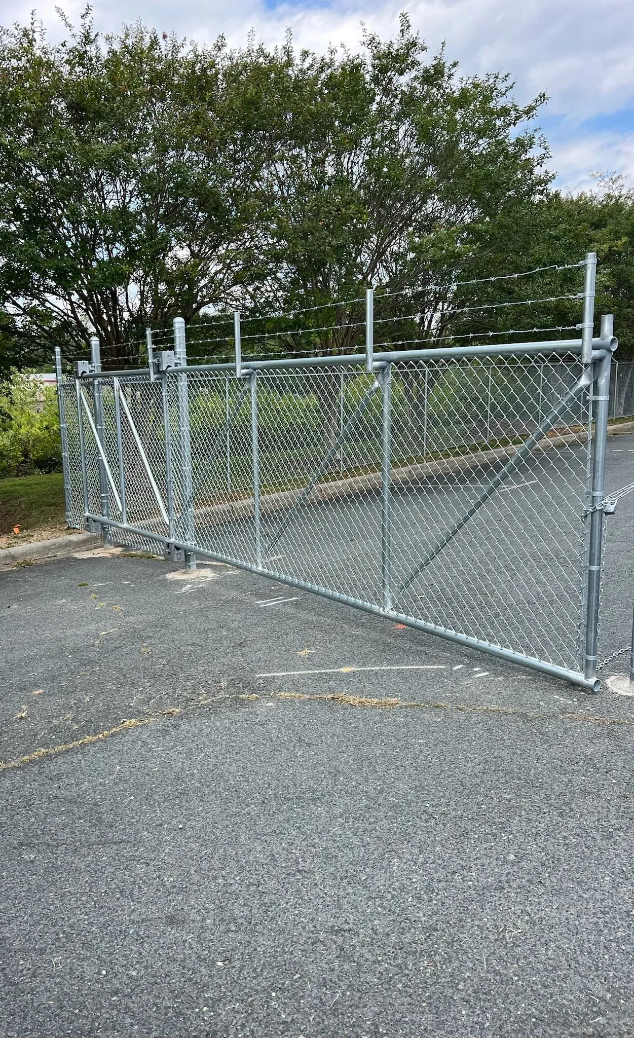 Chain-link fence with gate open on asphalt driveway, trees in background.