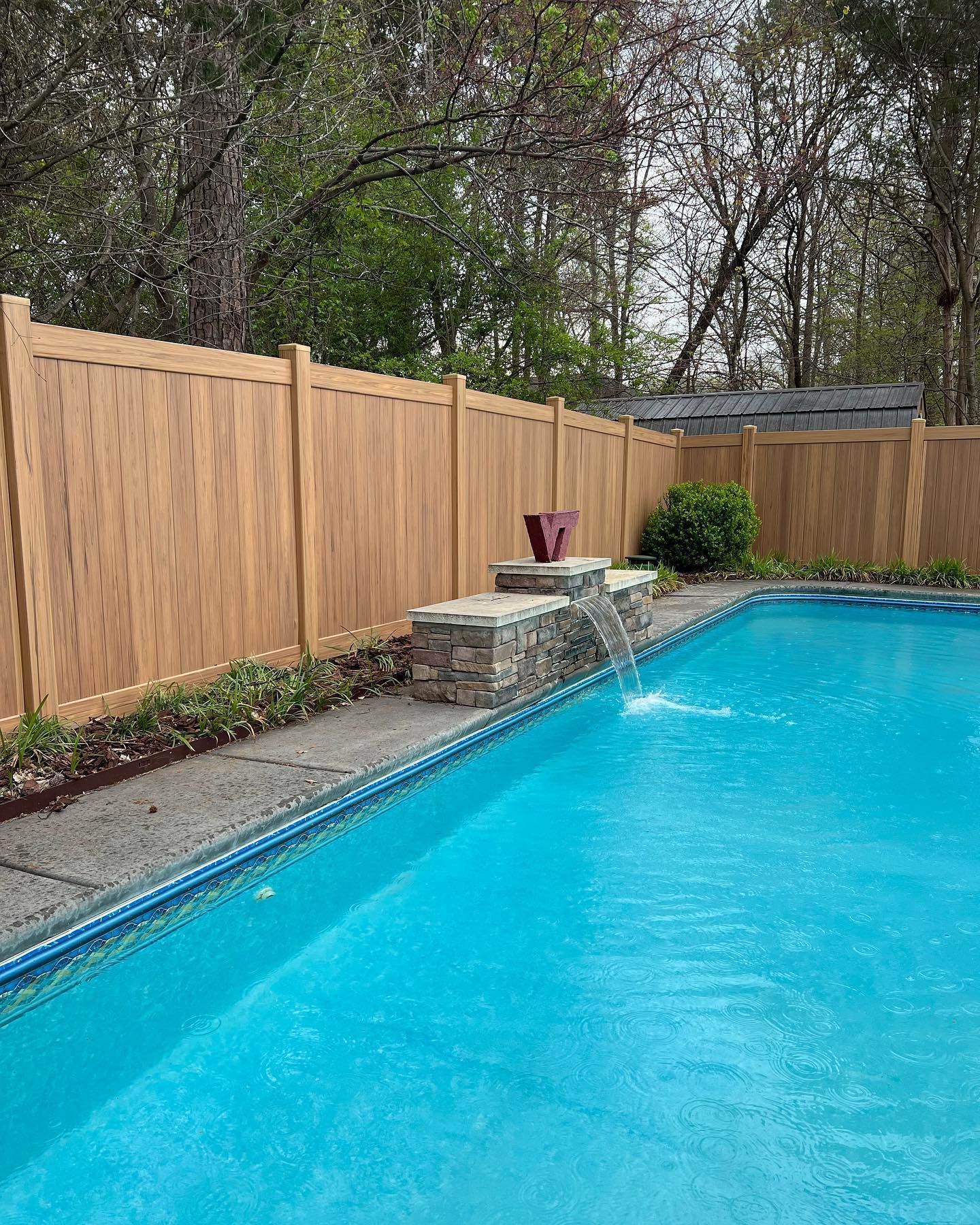 A backyard with a blue pool and a waterfall feature. A light brown fence borders the area.