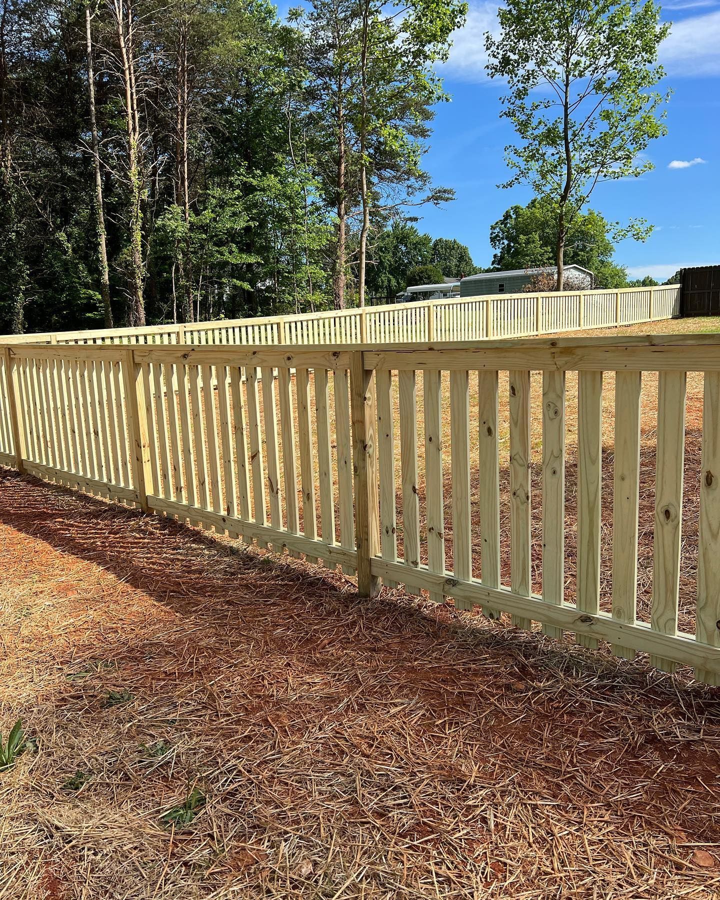 Wooden fence with vertical slats, on a grassy and dirt yard. Trees and blue sky in the background.