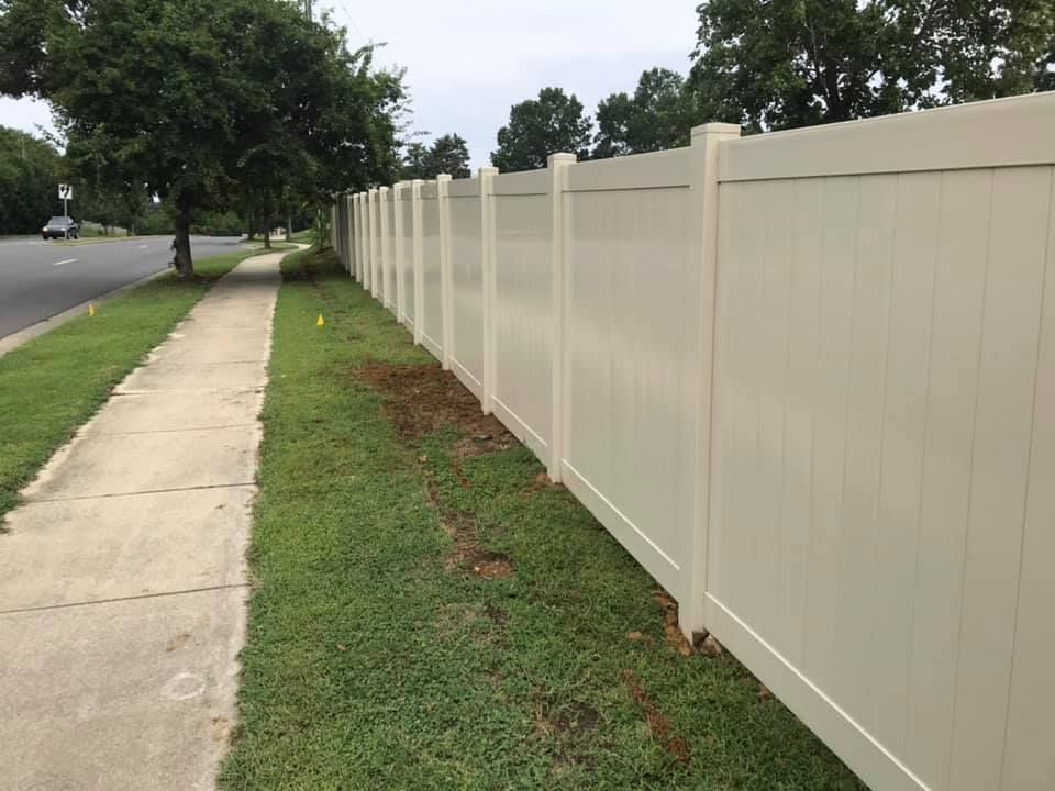 A beige vinyl fence runs along a sidewalk and grassy area next to a road, with trees in the background.