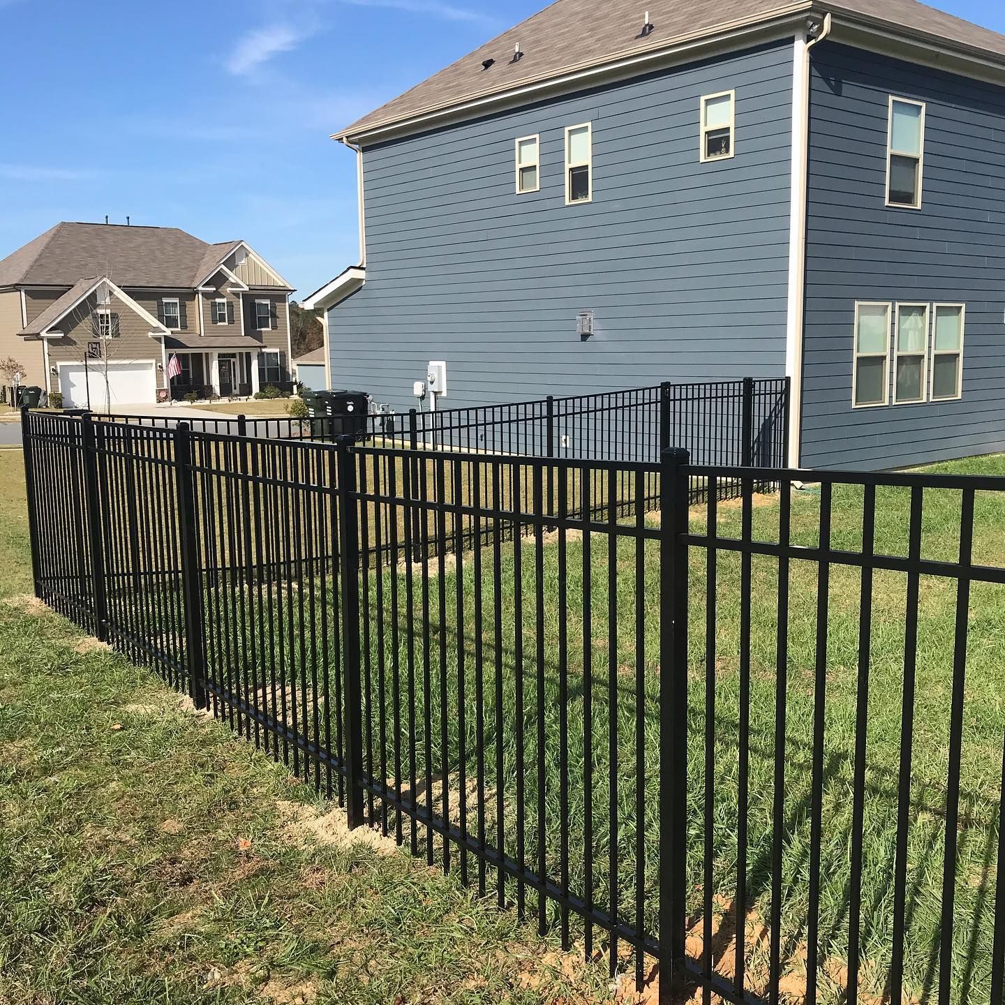 Black metal fence in a grassy yard, separating it from a blue house and other houses in the distance under a blue sky.