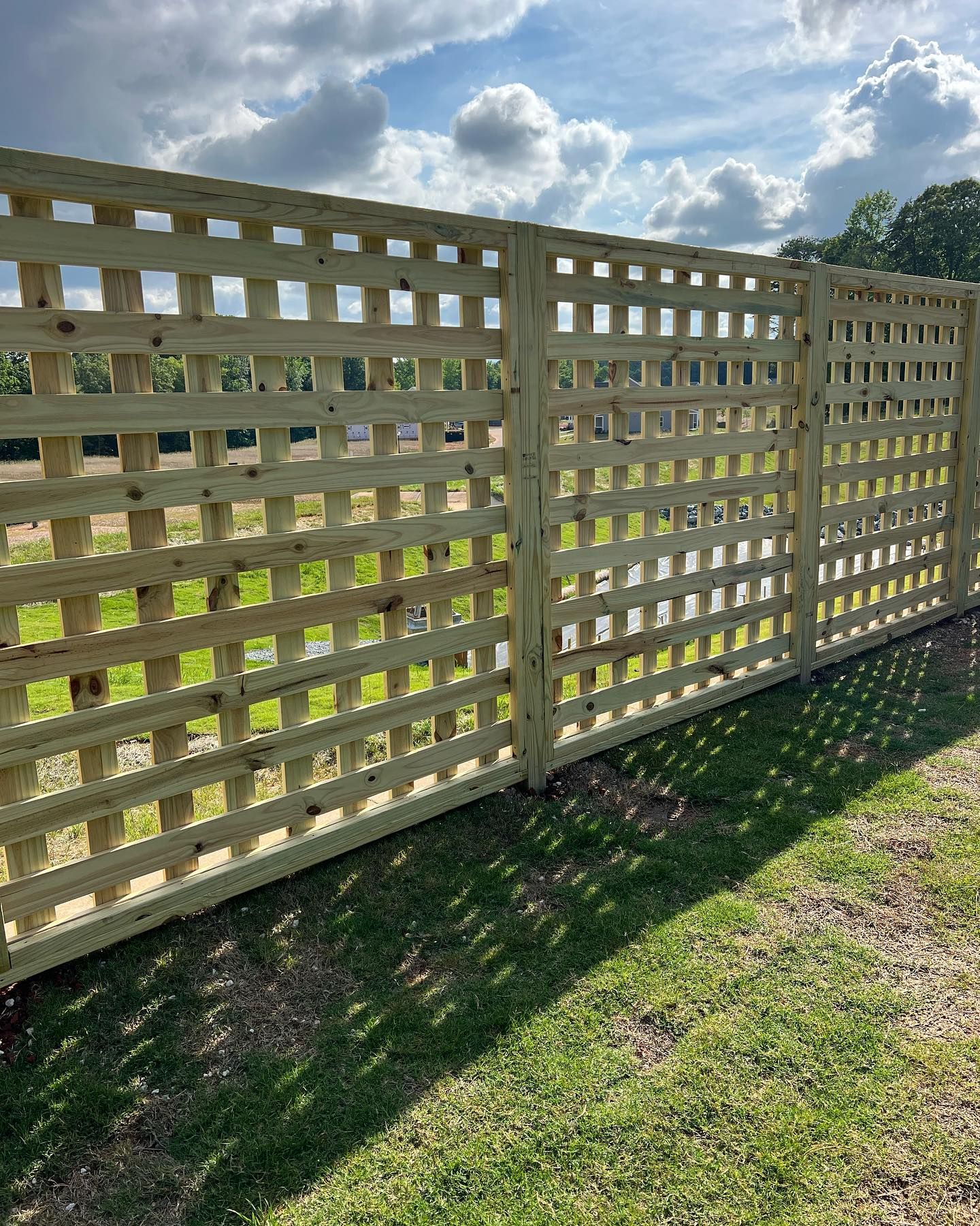 Lattice wooden fence in a grassy yard on a sunny day with clouds in the background.