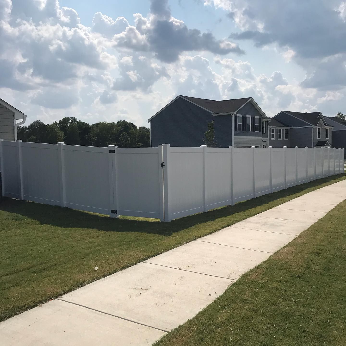 White privacy fence bordering a grassy lawn and a sidewalk, with houses in the background against a cloudy sky.
