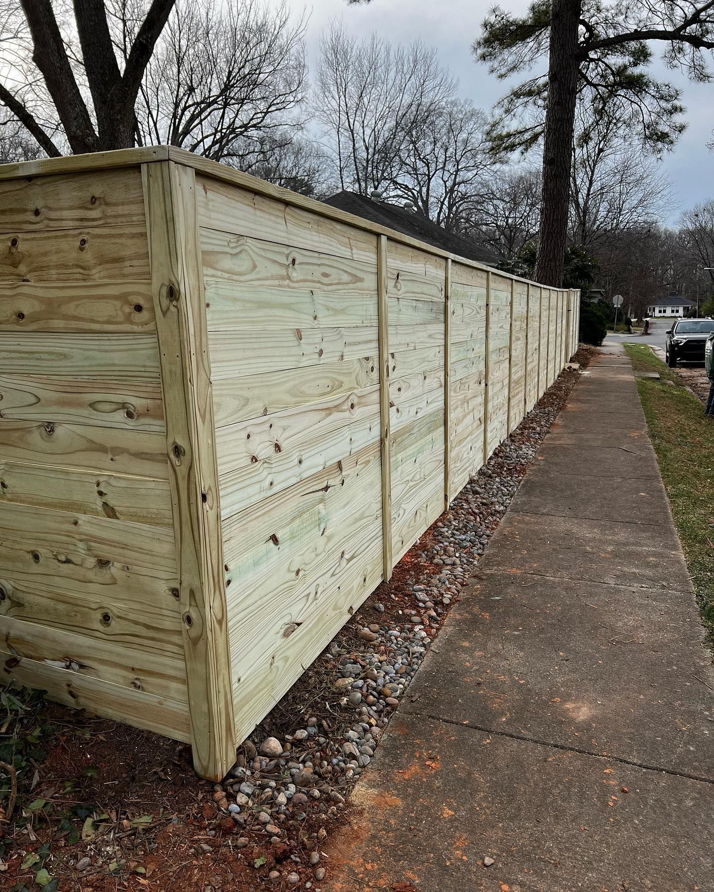 Wooden privacy fence along a sidewalk, with a bed of gravel at its base. The fence is constructed of horizontal planks.
