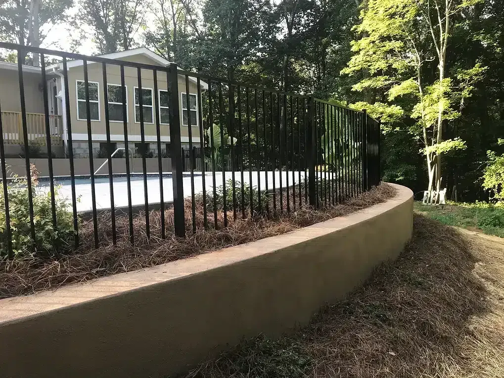 Black metal fence atop a low tan retaining wall borders a backyard with a pool and trees.
