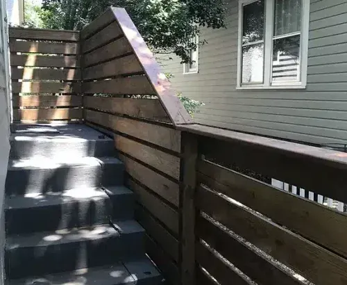 Exterior stairs with wooden railings next to a light-colored house with windows, under a tree.