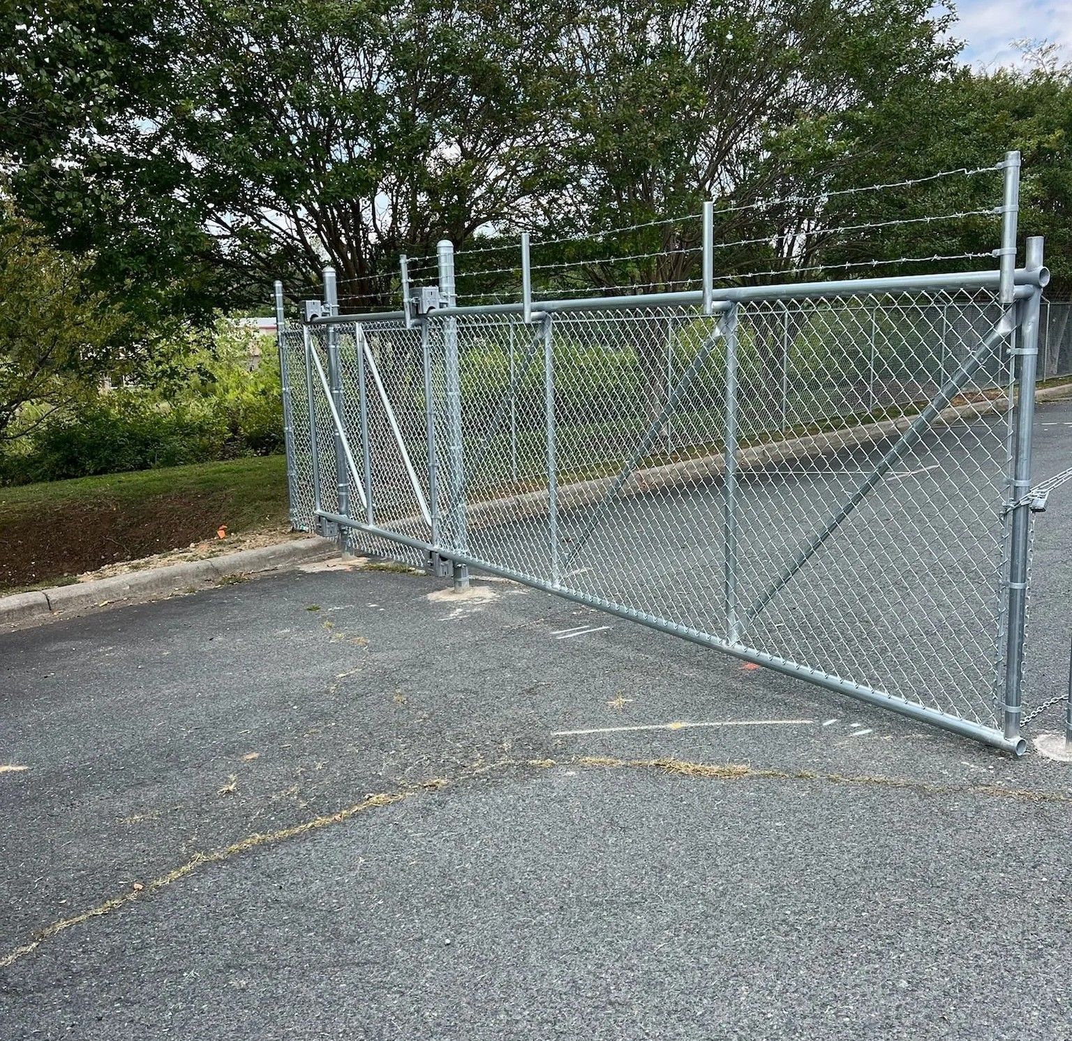 Chain-link fence with barbed wire, closing off an area, set on asphalt, trees in background.