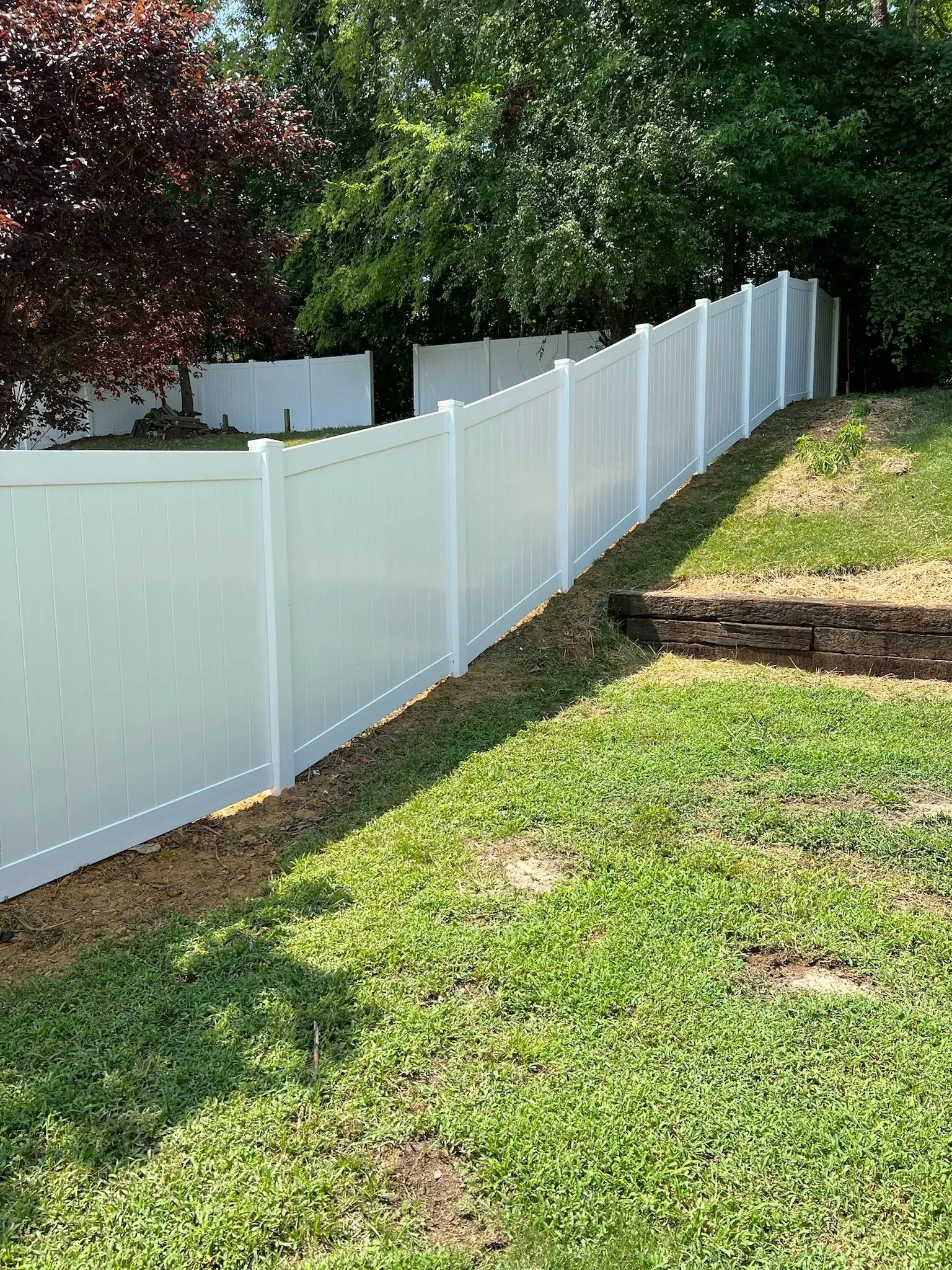 White vinyl fence on a grassy hill, separating properties with trees in the background.
