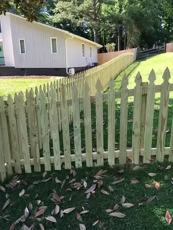 A wooden picket fence in front of a white house on a grassy hill.
