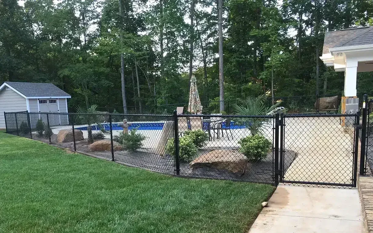 Black fence surrounds a backyard pool, garden, shed, and patio. Green grass and trees fill the background.