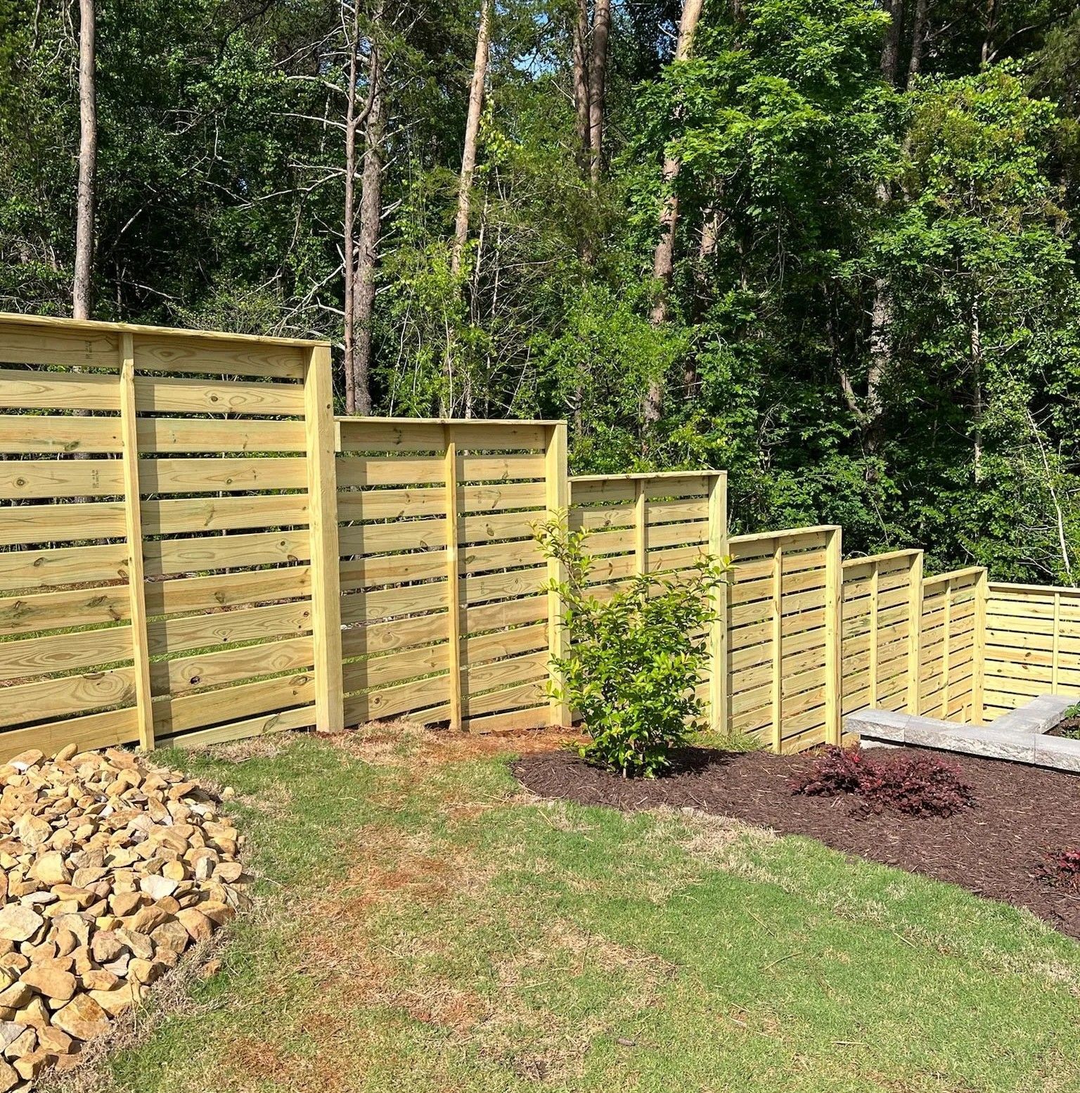 Wooden horizontal slat fence on a grassy yard bordering a wooded area.