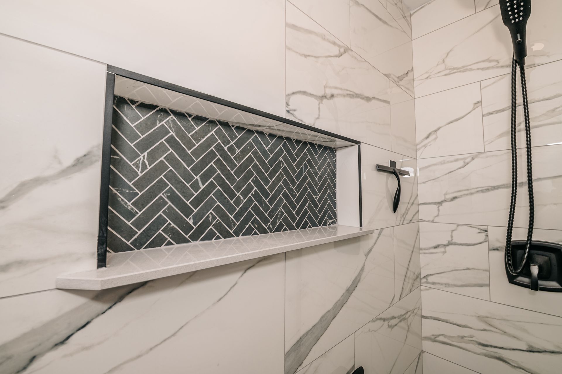 White tiled shower with a recessed shelf featuring gray herringbone tile and black fixtures.