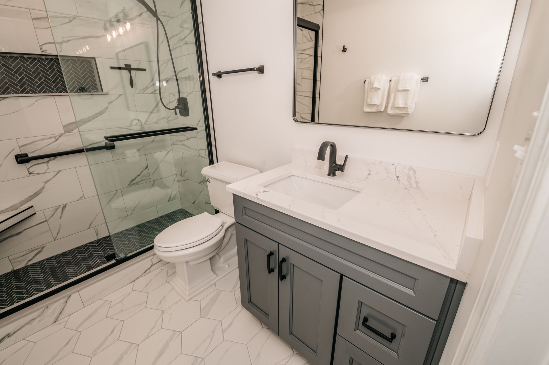 Modern bathroom with a white countertop vanity, toilet, glass shower with black trim, and large mirror. White and gray tile floor.