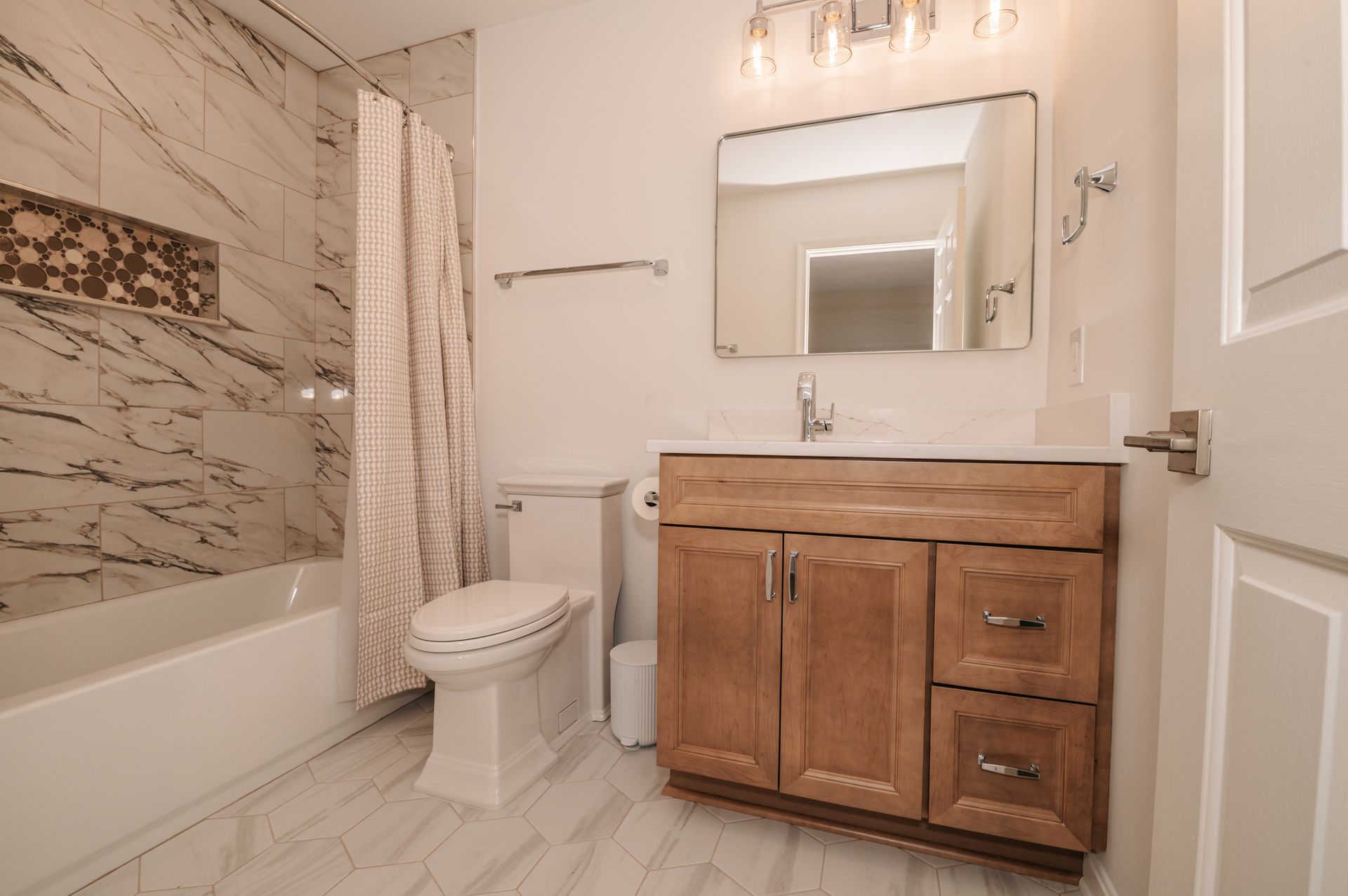 A modern bathroom with marble-look tile. Features a tub, toilet, vanity, and mirror. Soft lighting and a neutral color scheme.