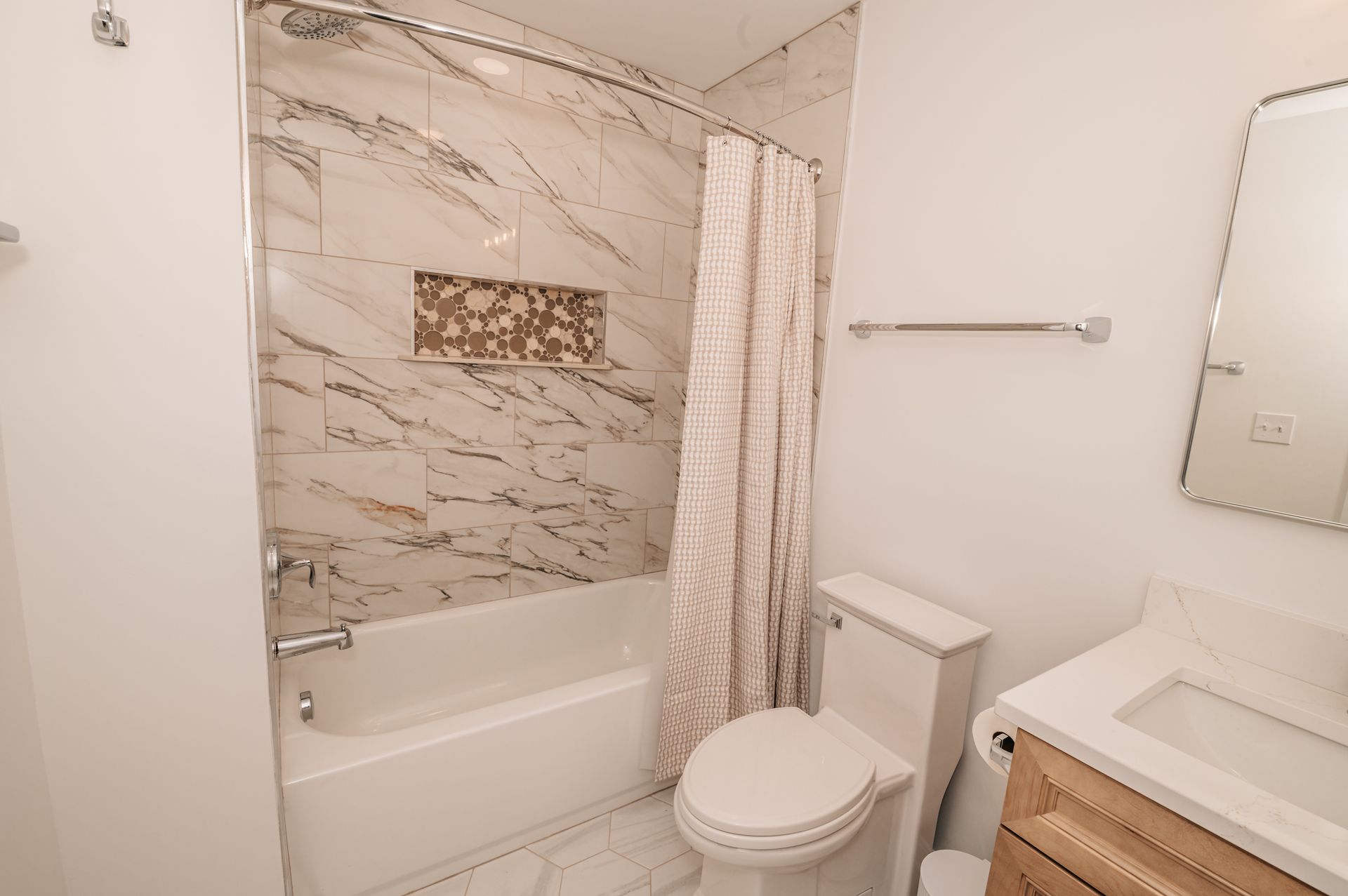 Small bathroom with a white tub and toilet, marble-patterned shower walls, a vanity with a mirror, and a beige shower curtain.