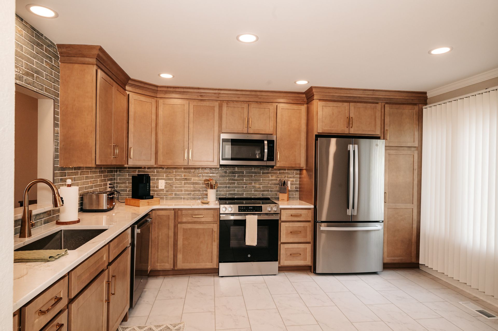 A modern kitchen featuring light brown cabinets, stainless steel appliances, and white countertops. A window is to the right with vertical blinds.