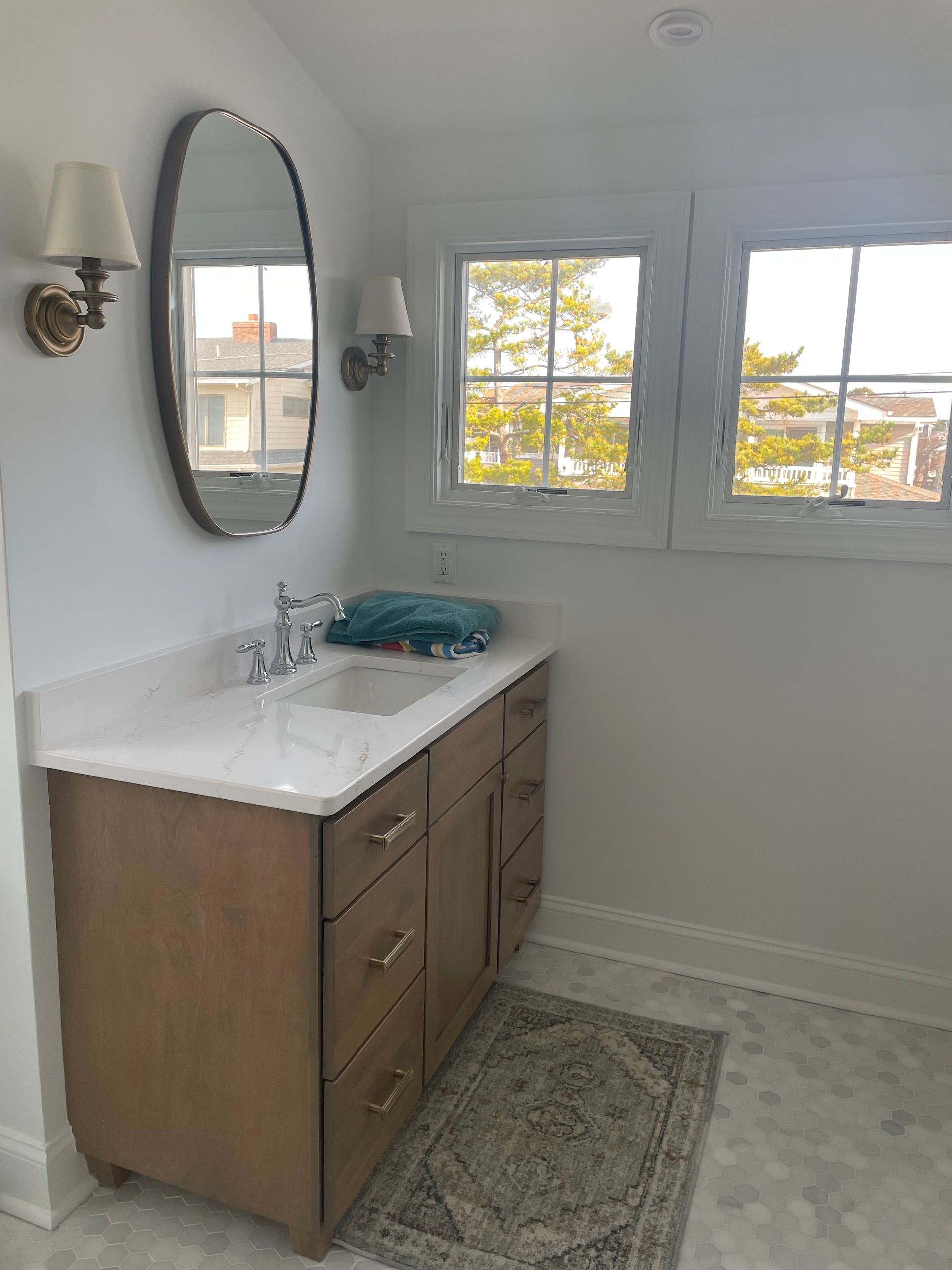 Bathroom with a wooden vanity, oval mirror, sconces, and a rug; a window overlooks a tree-lined view.
