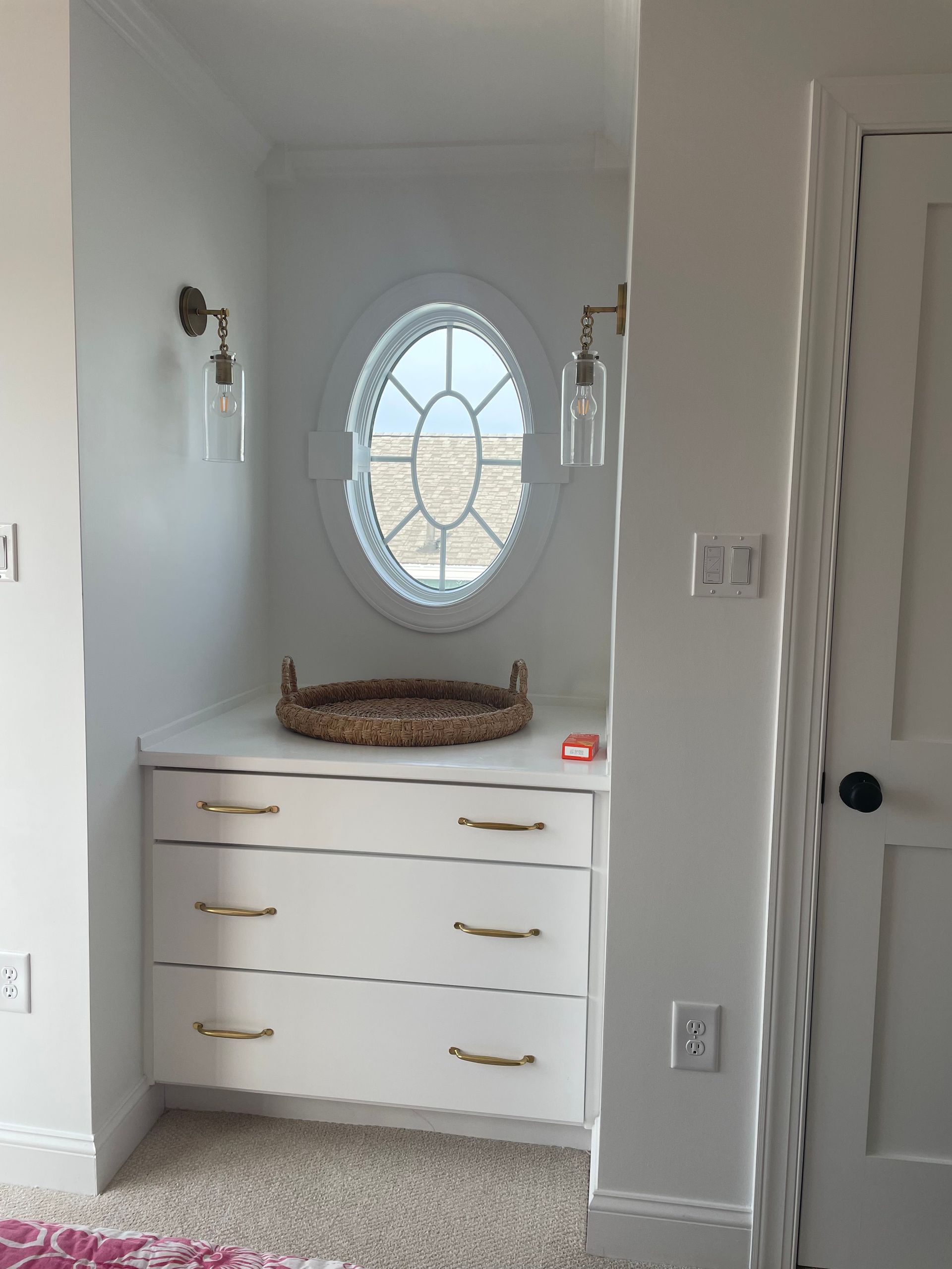 Built-in white dresser with brass handles, a decorative oval window, and sconce lighting. A woven tray sits on top.