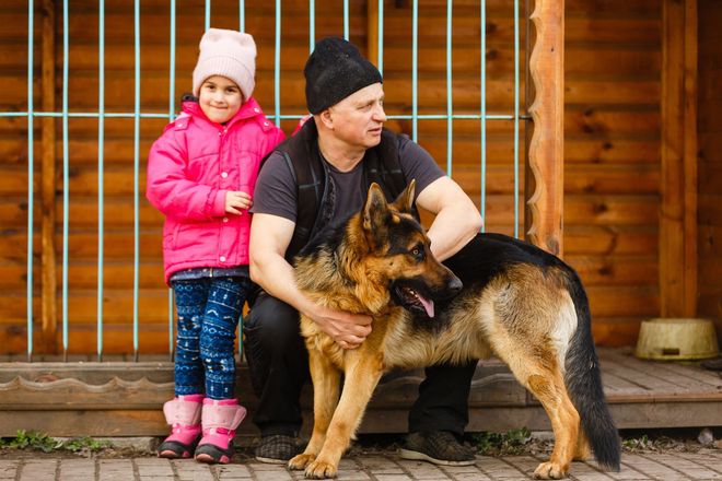 Man and girl with a German Shepherd dog in front of a wooden structure.