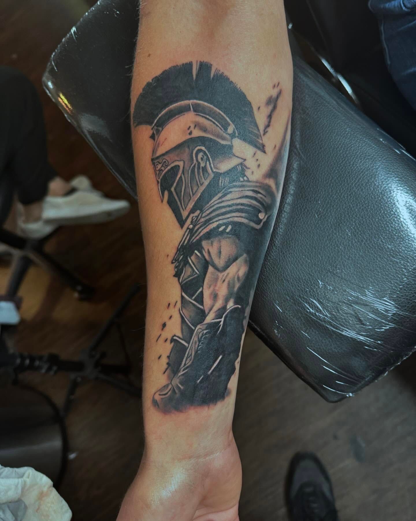 Forearm tattoo of a Spartan helmet on a person’s arm, shown on a black chair.
